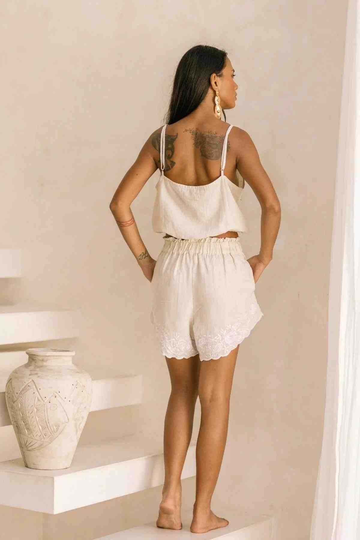 A woman with long dark hair stands on white steps, facing away. She is dressed in the Wonder Sleeper Set from Myrah Penaloza, a beige sleeveless top and matching shorts featuring lace trim, reminiscent of delicate sleepwear. Nearby rests a large white vase, while a light-colored wall provides a serene backdrop.