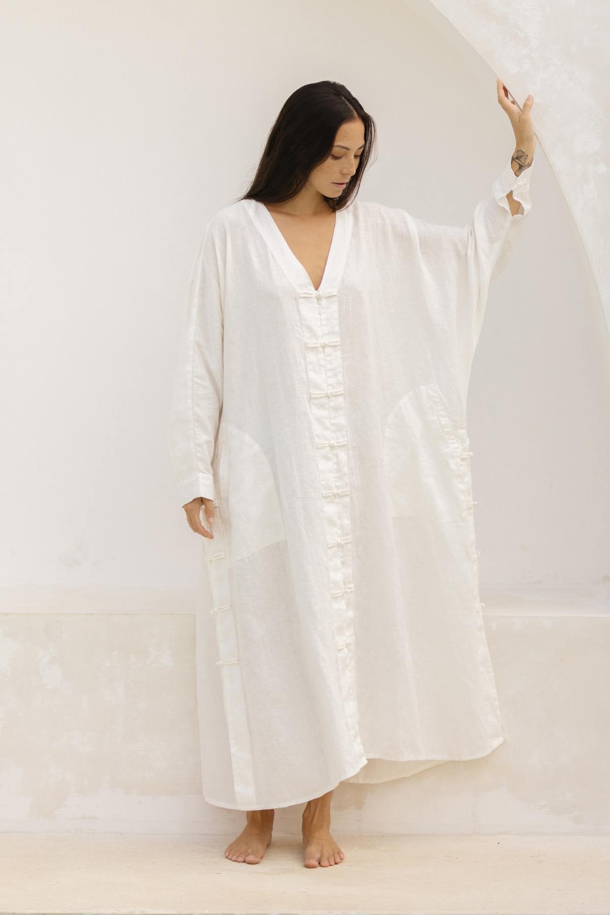 A person with long dark hair stands barefoot inside a minimalistic setting. They are wearing a loose-fitting, long white dress with a V-neck and long sleeves, resembling the Ancient Chajin Jacket from Myrah Penaloza. One hand is resting against a wall while the other hangs loosely by their side.