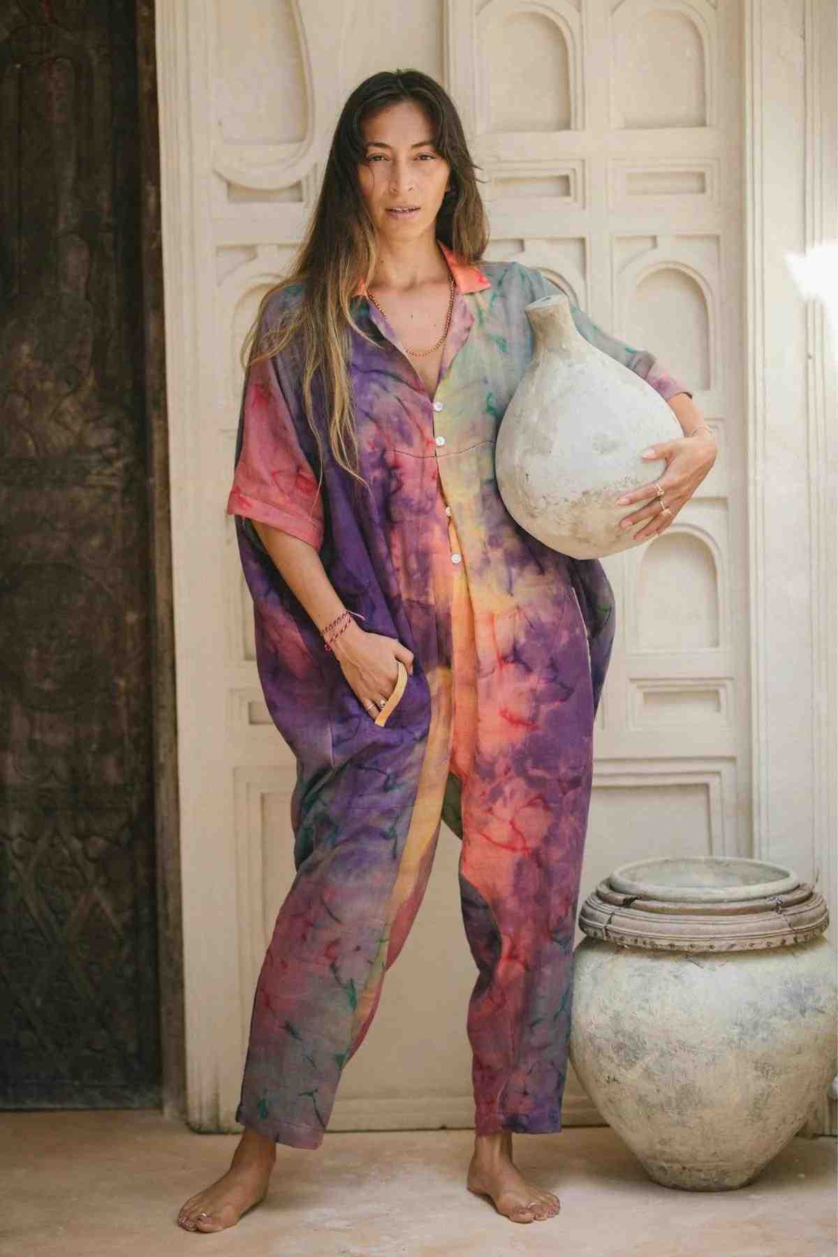 A person stands barefoot in a doorway, dressed in the vibrant Sat Torri Rainbeau Playsuit from Myrah Penaloza with its retro 1980s design and handcrafted feel, holding a large clay vessel. Another similar vessel rests on the ground beside them. The background features intricate wall designs and a partially open dark wooden door.