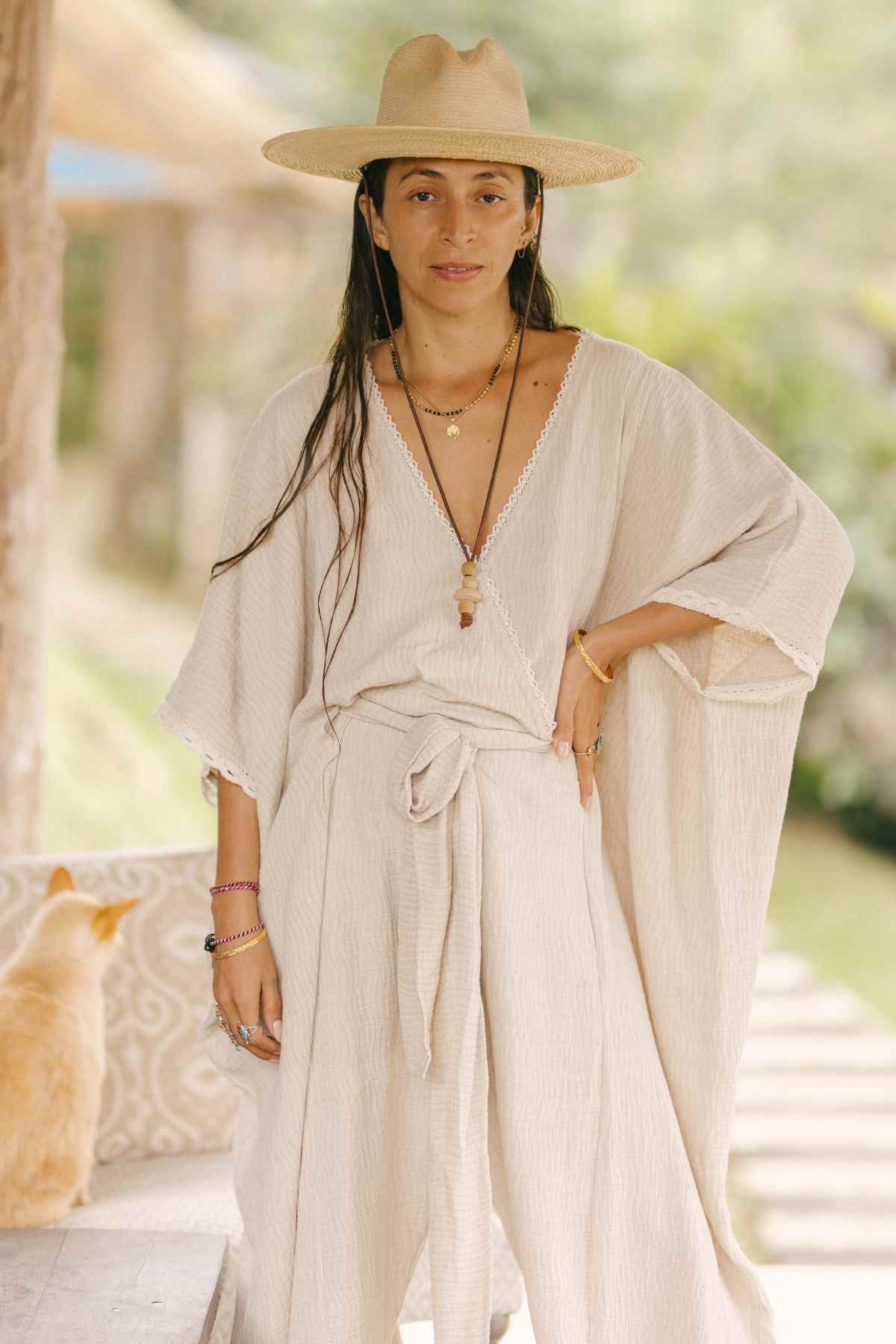 A person wearing a flowing Limited Edition Hand Embroidered River Playsuit by Myrah Penaloza, paired with billowy sleeves and a wide-brimmed hat, stands outdoors. They have layered necklaces and bracelets, while a peach-colored cat sits nearby on a ledge. The background is blurred greenery, suggesting a tranquil setting.