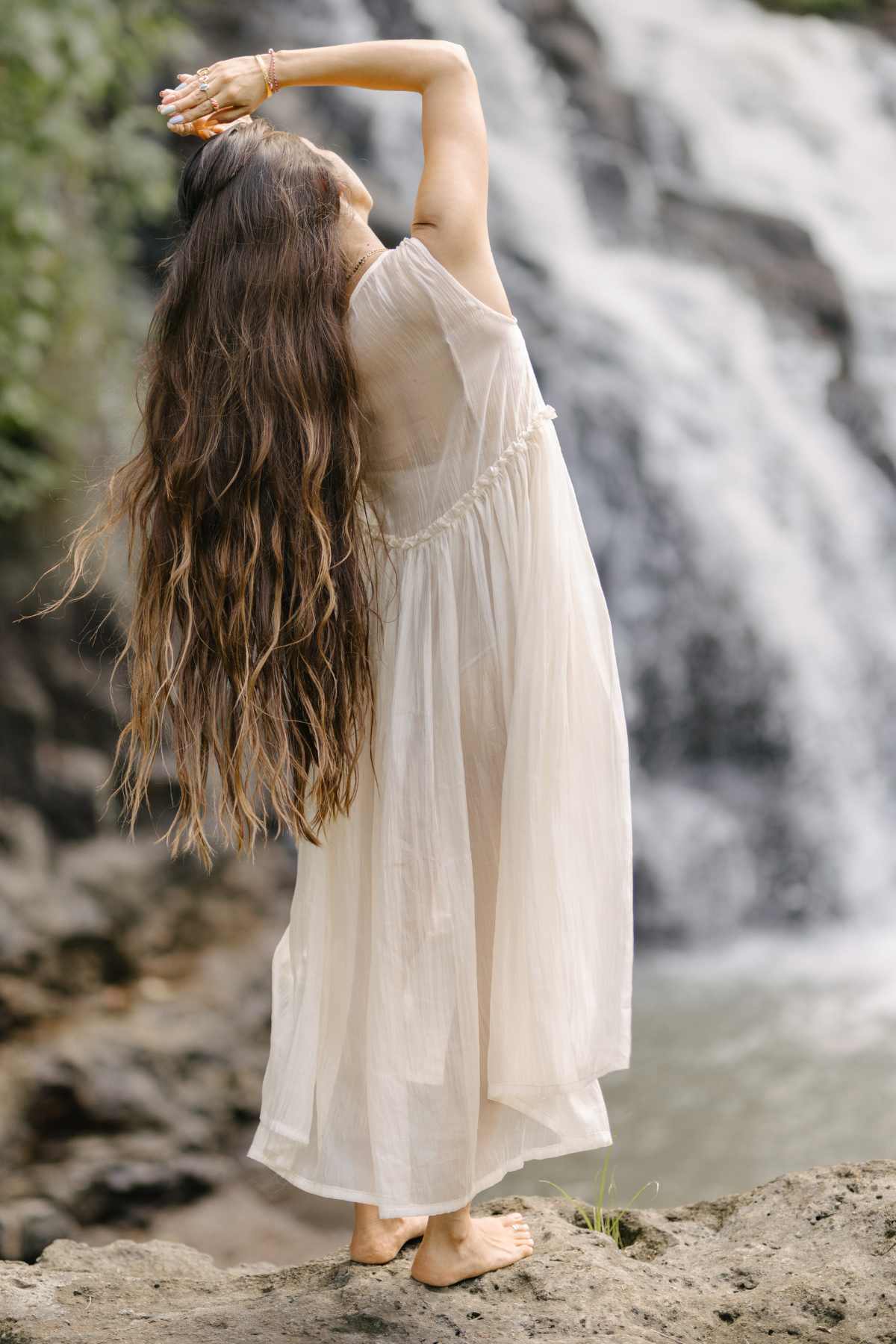 A woman with long, wavy hair and wearing the Dolly Night Gown (Sheer, 100% Chiffon Silk) by Myrah Penaloza stands barefoot on a rock near a waterfall. She is stretching with her arms overhead, surrounded by natural scenery.