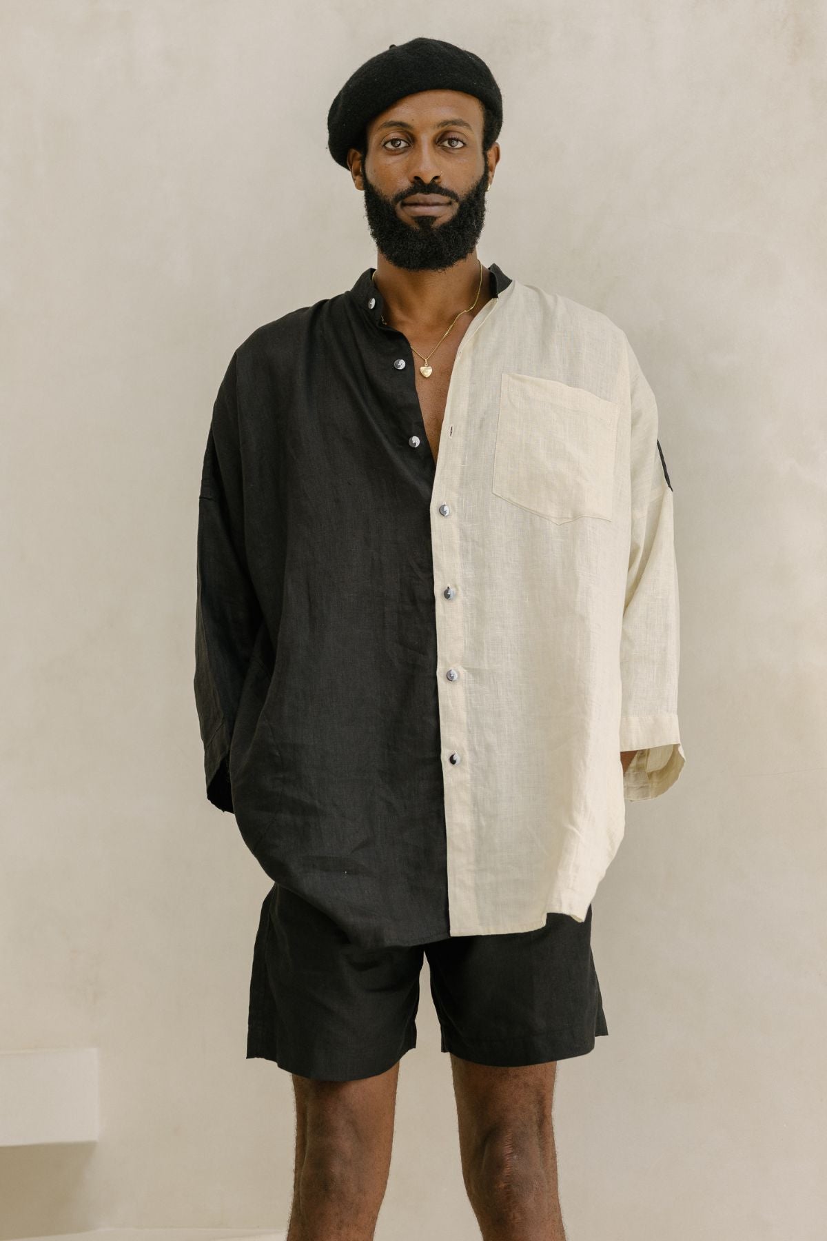 A person dressed in the Camisa del Novio Mens Linen Top by Myrah Penaloza, featuring a stone-washed two-toned black and white design, paired with black shorts, stands against a neutral background. They complete their look with a black beret and have a beard.