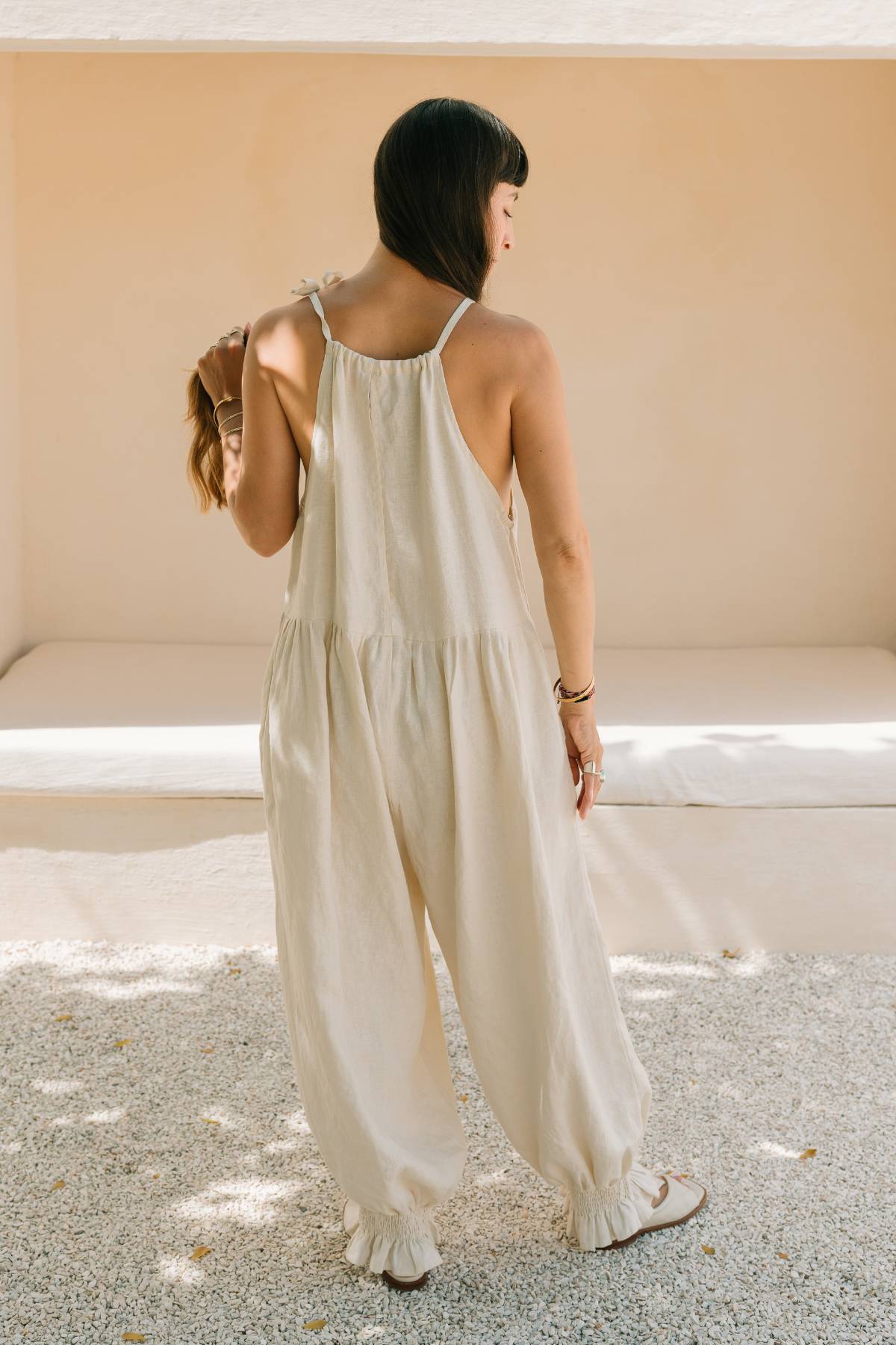 Back view of a woman wearing the Monaya Playsuit by Myrah Penaloza in Moonlight stone washed linen. Oversized, full length silhouette with tie shoulder straps and a tiered back yoke, standing on a gravel courtyard.