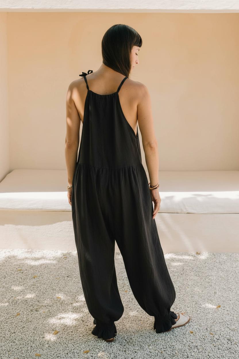 Back view of a woman wearing the Monaya Playsuit by Myrah Penaloza in Dark Moon black stone washed linen. Oversized, full length silhouette with tie shoulder straps and a tiered back yoke, standing on a gravel courtyard.