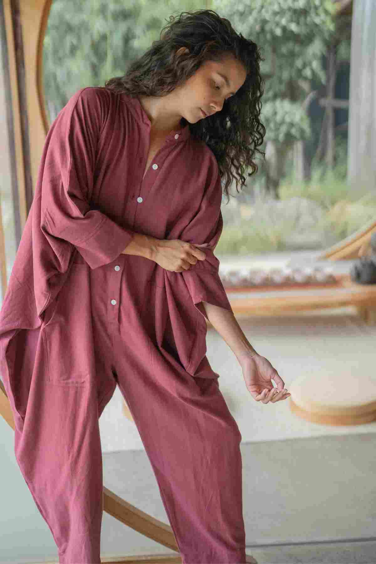 A person with wavy hair is wearing a loose-fitting, long-sleeve, button-up jumpsuit in a reddish color from the limited edition Miracle Playsuit 2.0 (Karama Collection) by Myrah Penaloza. Standing indoors in front of a large window, they adjust their sleeve and look down. The background showcases greenery and modern wooden elements.