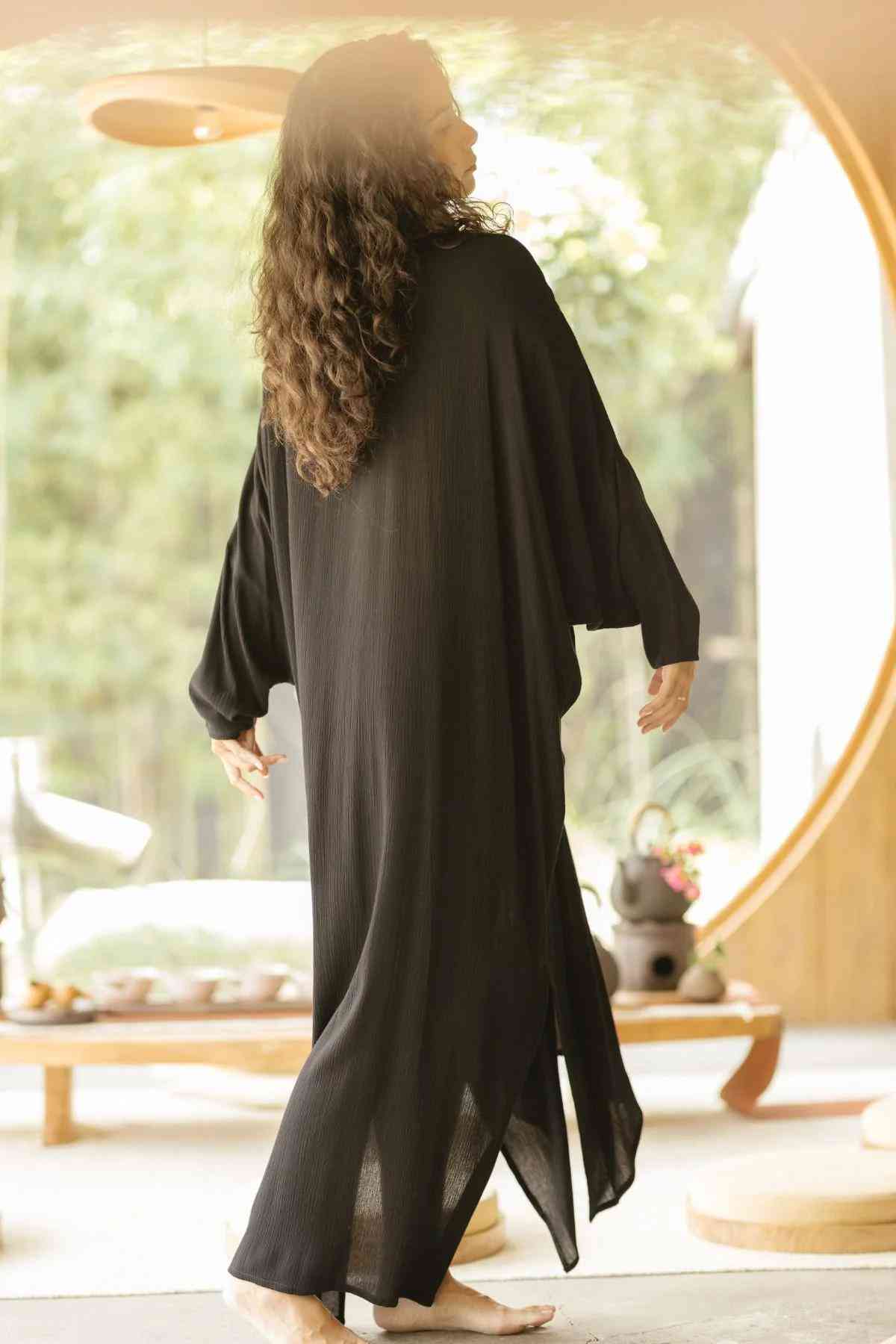 A person with long, wavy hair stands barefoot on a shaded patio, wearing the flowing Bamboo Rayon Virgo Rising Gown by Myrah Penaloza. They are facing away from the camera, with sunlight filtering through an opening in the background, highlighting a lush, green outdoor setting.