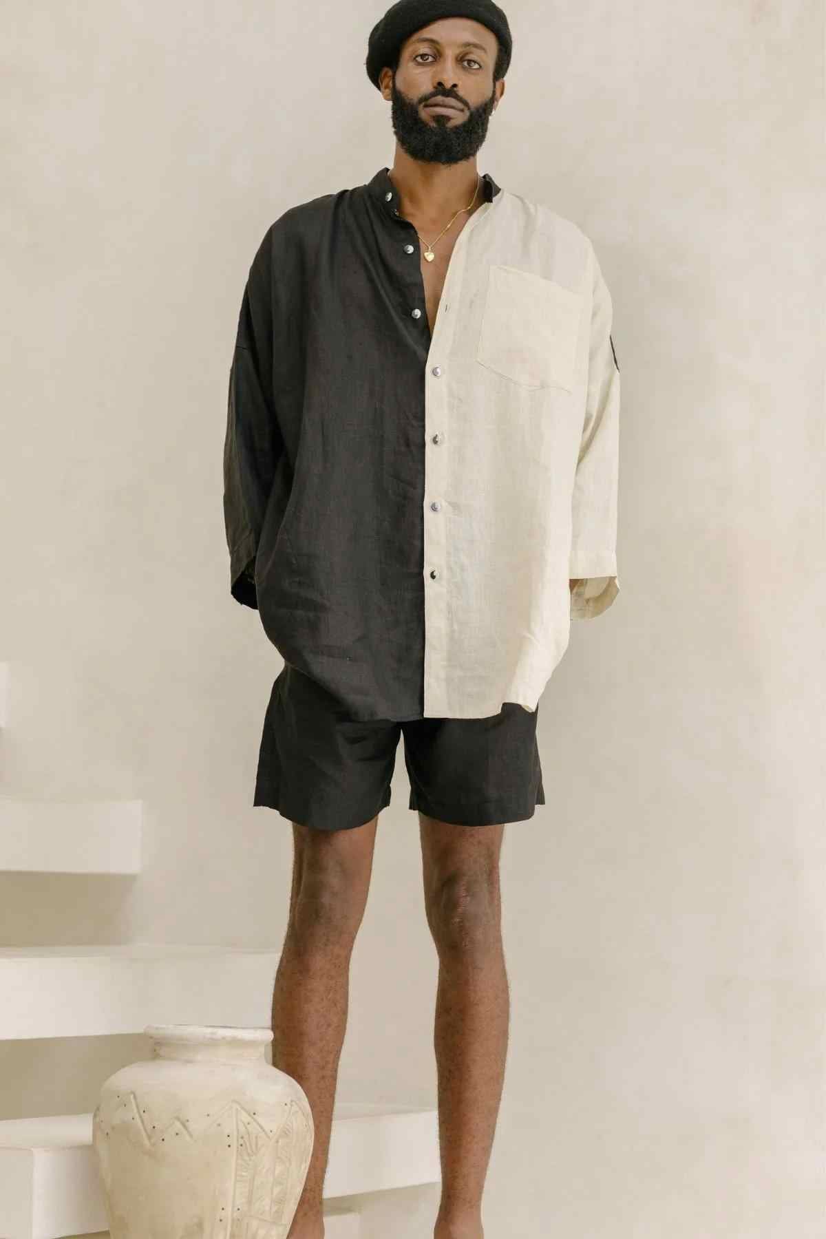 A man stands on white steps, wearing the Unisex Yin Yang Linen Camisa Del Novio Set by Myrah Penaloza, which includes a handmade half-black, half-beige long-sleeved shirt paired with black shorts and a black beret. He has a beard and is barefoot. A large decorative vase sits at the foot of the steps. The background is plain and neutral in color.