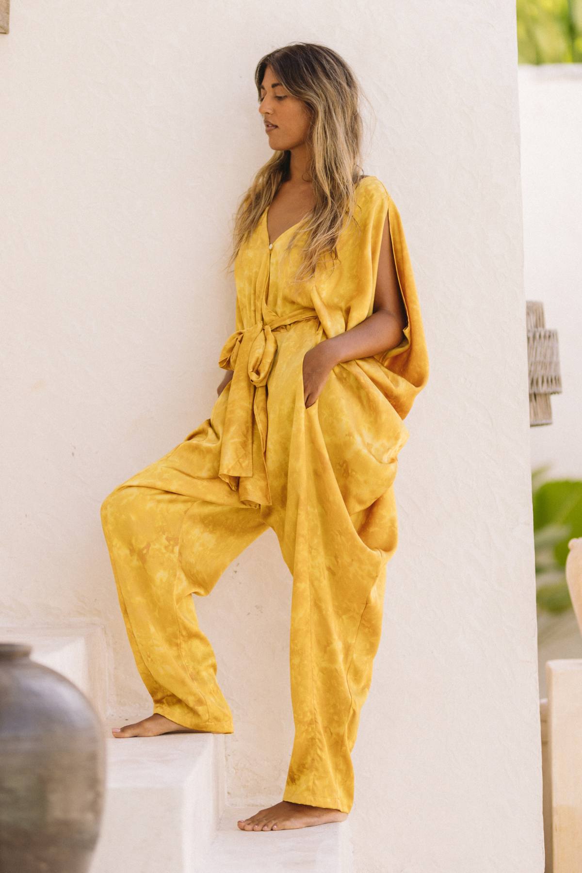 A woman with long hair stands on white stairs, leaning against a wall. She is wearing a loose-fitting Kuan Yin Playsuit by Myrah Penaloza, made to order from 100% botanically dyed turmeric gold silk. The playsuit features short sleeves and a waist tie. One hand rests in her pocket as large pottery and greenery adorn the background.
