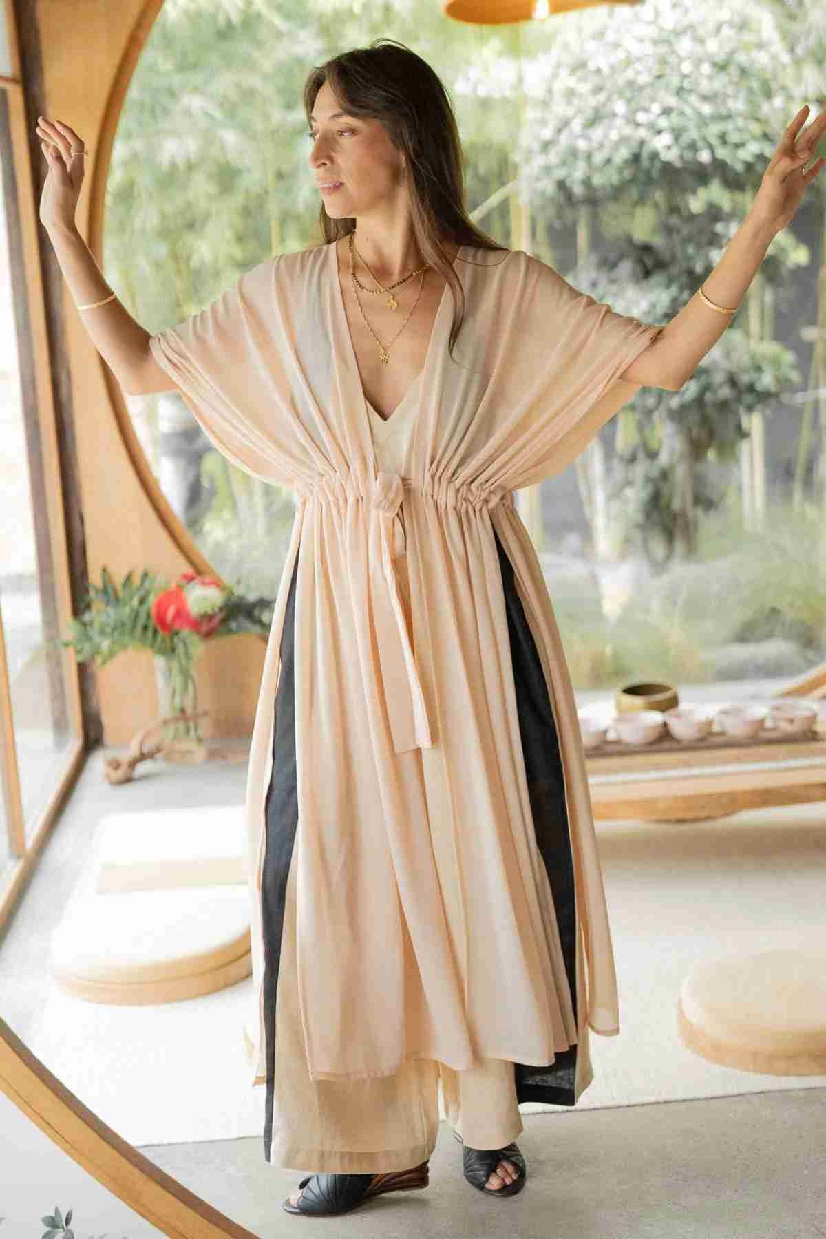 A woman stands in a relaxed pose with open arms, wearing a flowing, light-colored dress with a deep V-neck and cinched waist. The Madelin Sheer Chiffon Long Cover by Myrah Penaloza accentuates her sheer brilliance. She is in a peaceful, minimalist room with large circular windows and natural greenery in the background.