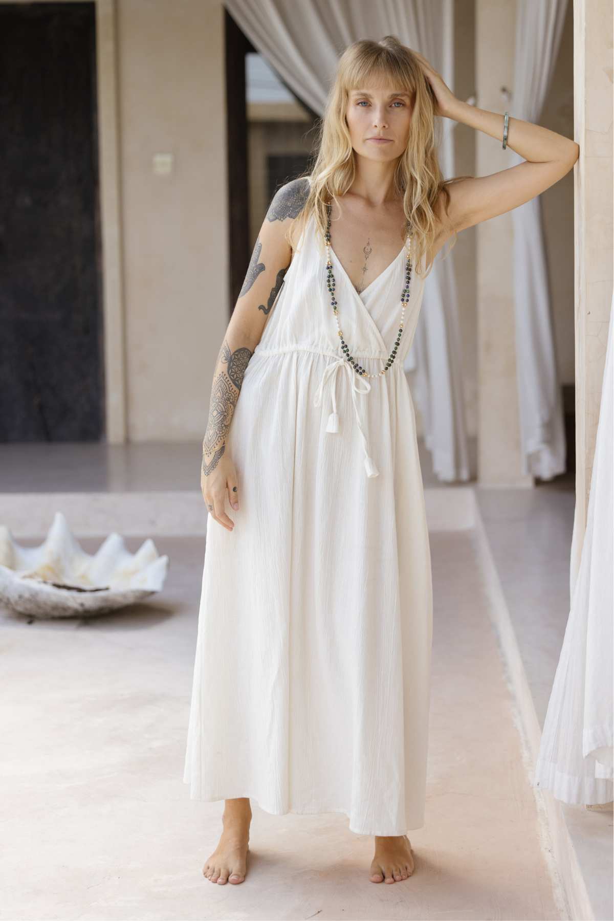A person with long blonde hair stands barefoot on a smooth floor, effortlessly elegant in a flowing white Dora Slip Dress by Myrah Penaloza, complemented by beaded necklaces. Their right arm is adorned with tattoos as they stand in a light-colored indoor space with draped curtains.