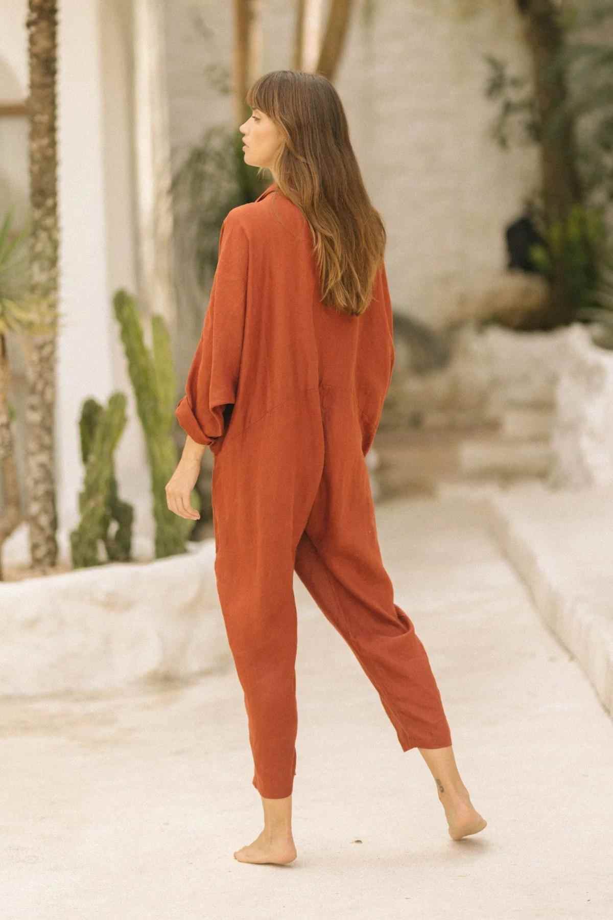 A person with long brown hair stands barefoot on a smooth, pale surface. They are wearing an oversized Linen Amma Playsuit by Myrah Penaloza, made of linen/cotton crinkle material and are viewed from the back. The setting includes white walls, green plants, and a minimalistic, natural aesthetic.
