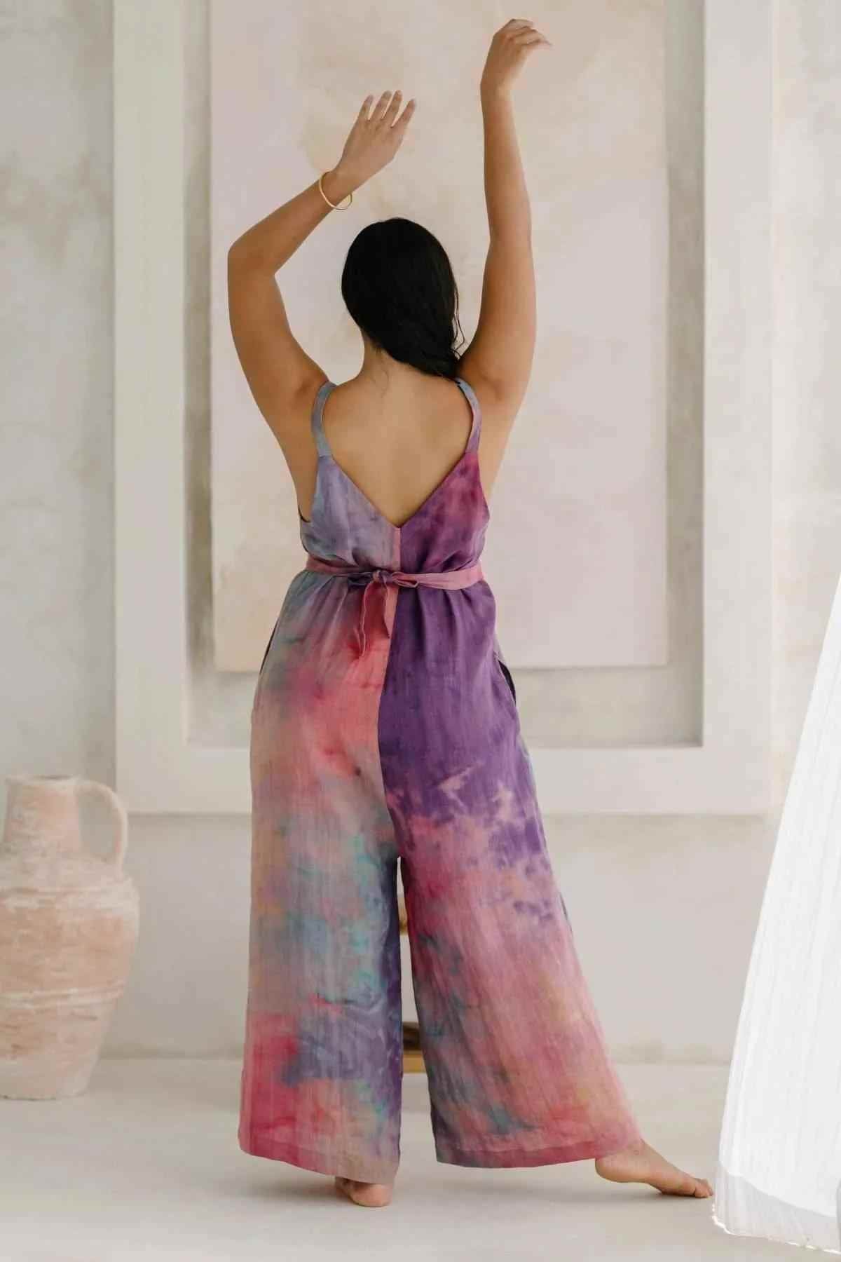 A person with dark hair in a ponytail stands with their back to the camera, arms raised. They are wearing an elegant Rainbeau Linen Kundalini Playsuit by Myrah Penaloza, featuring shades of purple, pink, and blue. An off-white urn is placed on the left, and a soft light illuminates the scene.