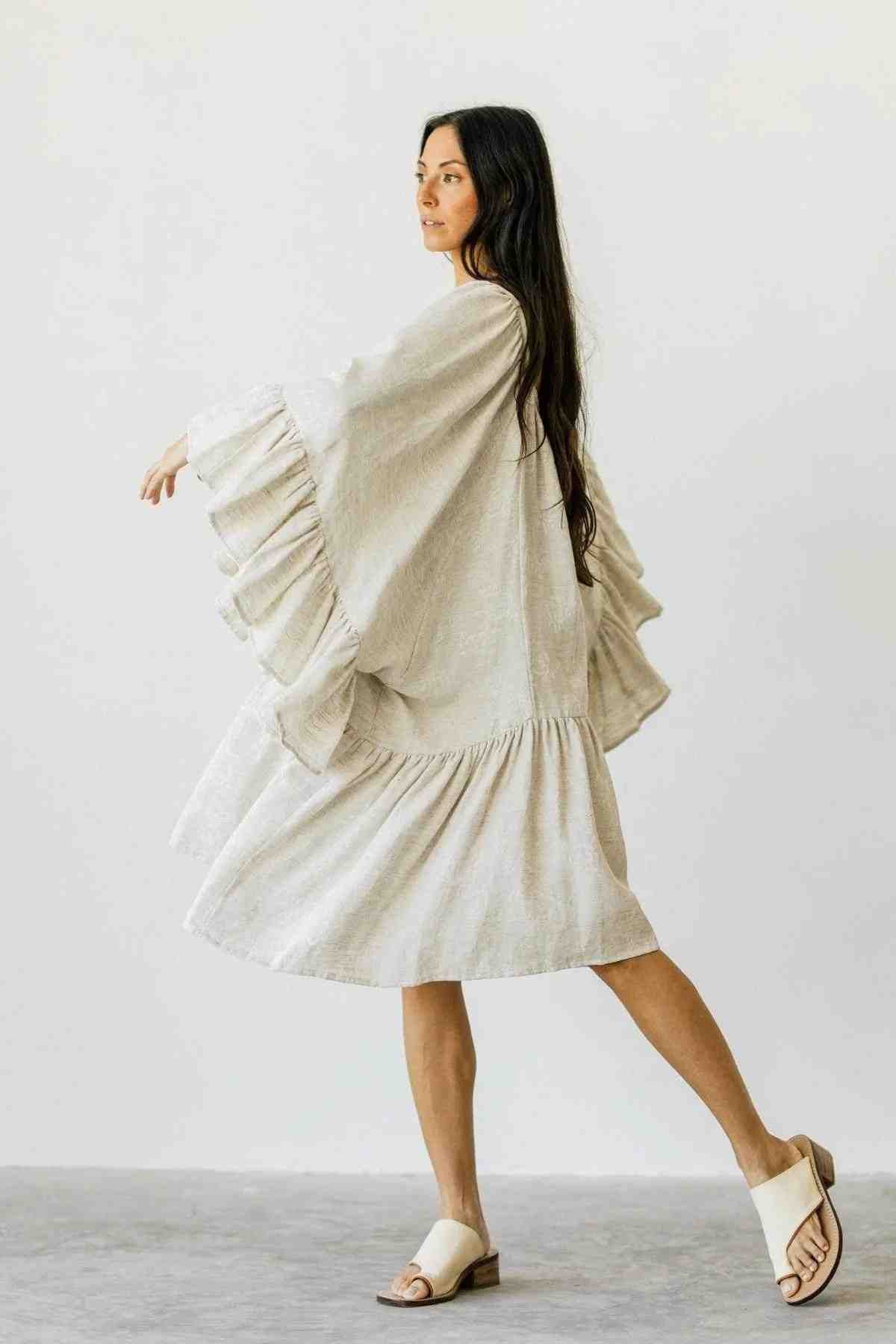A person with long dark hair is gracefully walking, dressed in a romantic spring/summer Linen Brida Gown Short by Myrah Penaloza, featuring ruffled sleeves. They're also wearing beige sandals. The plain light background enhances the minimalist and airy atmosphere, perfectly complementing the gown's charm.