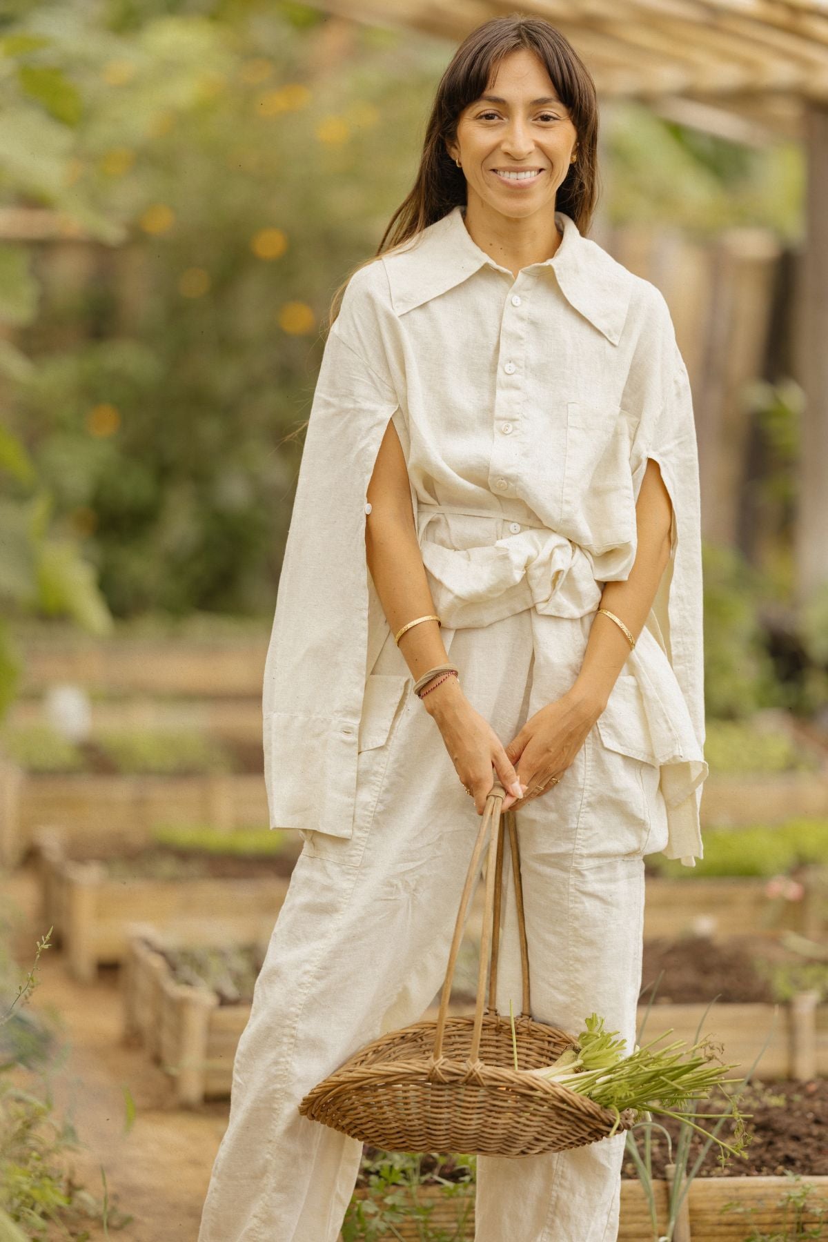 A woman stands smiling in a garden, dressed in the Sutra Linen Set by Myrah Penaloza. The light-colored, loose-fitting outfit with a large collar is crafted from versatile, lightweight linen. She holds a wicker basket filled with green plants, surrounded by garden beds and lush greenery.
