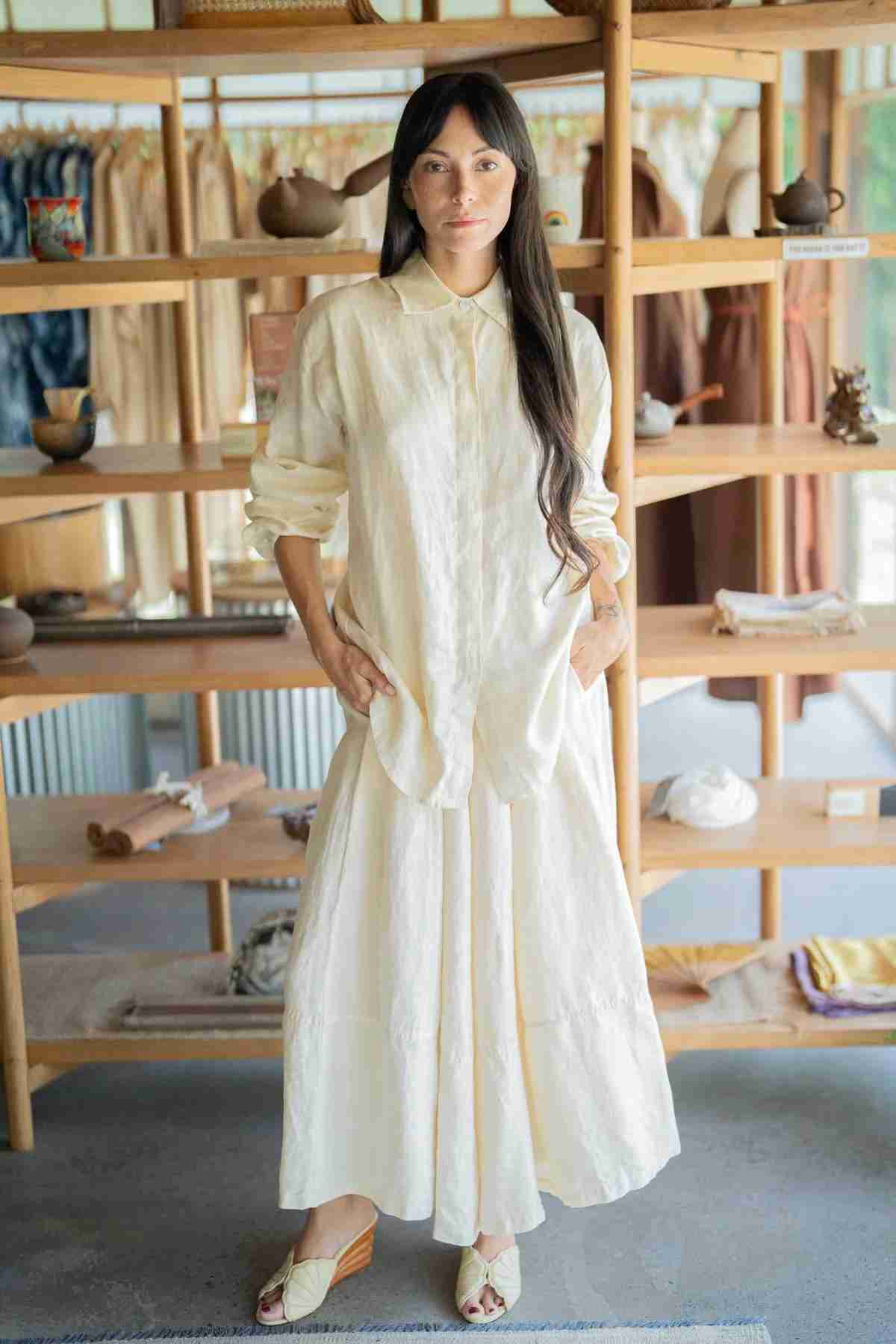 A woman with long dark hair stands in front of a wooden shelving unit filled with various household items. She is wearing the Pantalon Linen Pants by Myrah Penaloza, which have a wide-legged and oversize fit, along with pointed shoes, her hands casually in her pockets. The background features shelves lined with folded fabrics.