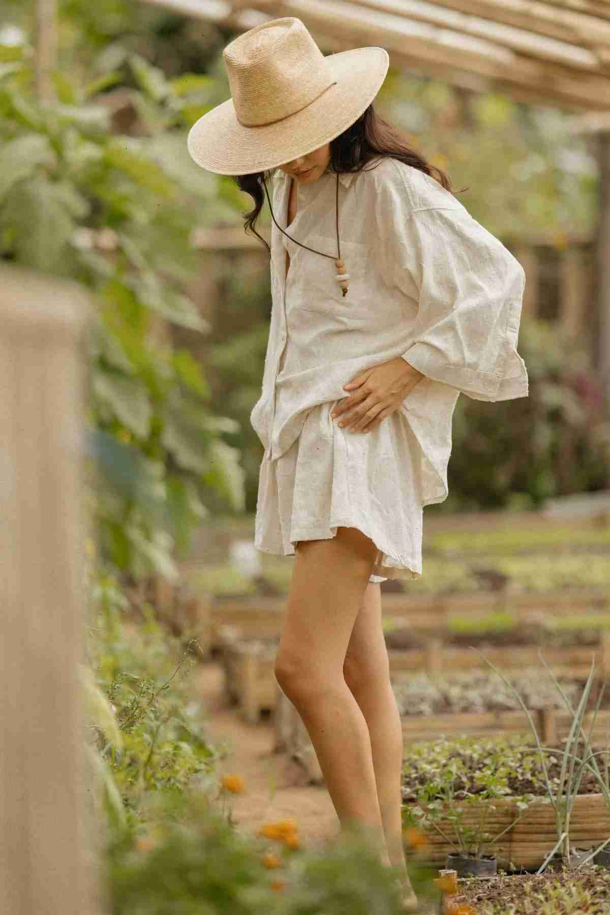 Dressed in the Linen Nidra Button Down Set by Myrah Penaloza, a woman stands gracefully in a garden. With her gaze lowered and a wide-brimmed hat shading her face, she is enveloped by lush greenery and raised garden beds. The calm, sun-drenched setting beautifully embodies nature's elegance.