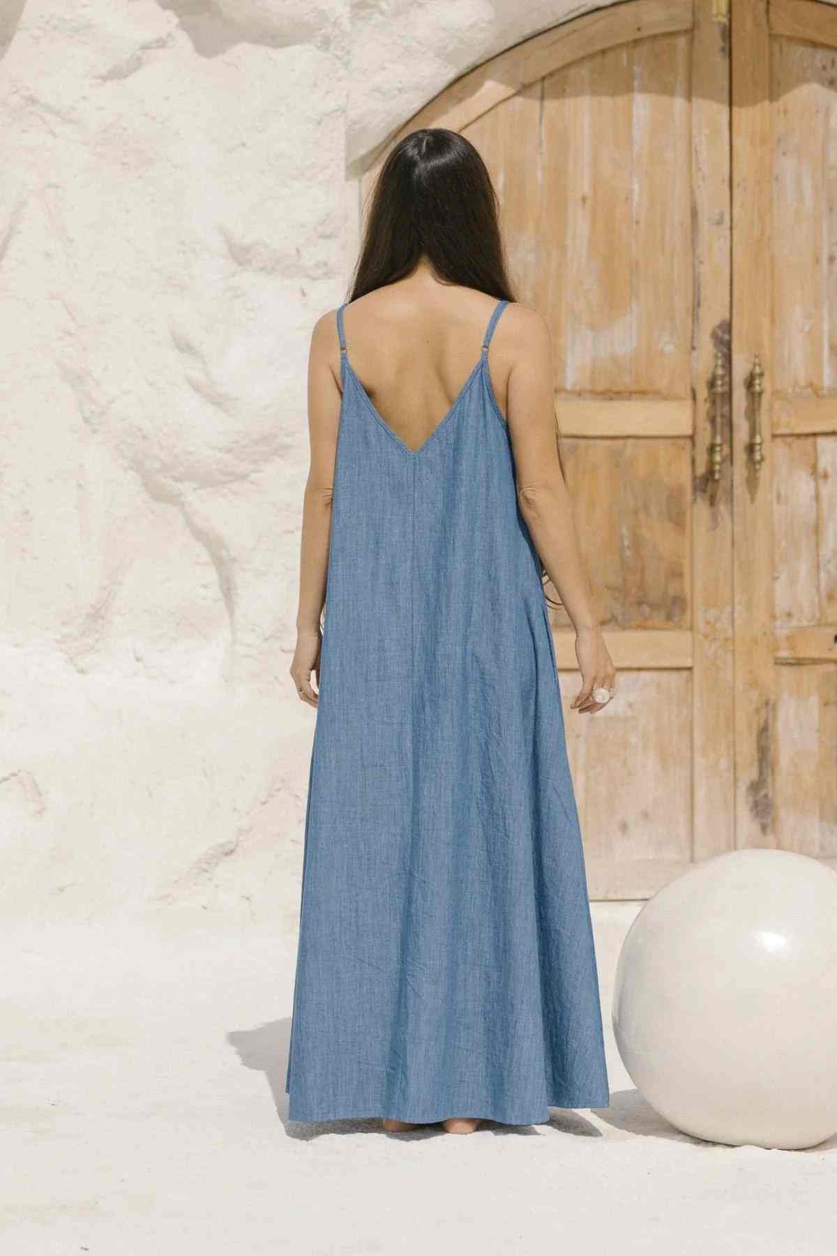 A woman with long dark hair stands facing a rustic wooden door, adorned in the exquisite Kundalini Denim Dress from Myrah Penaloza, featuring a sleeveless design and a V-shaped back. The minimalist, sunlit scene is enhanced by a white round object on the ground beside her, elevating the serene ambiance.