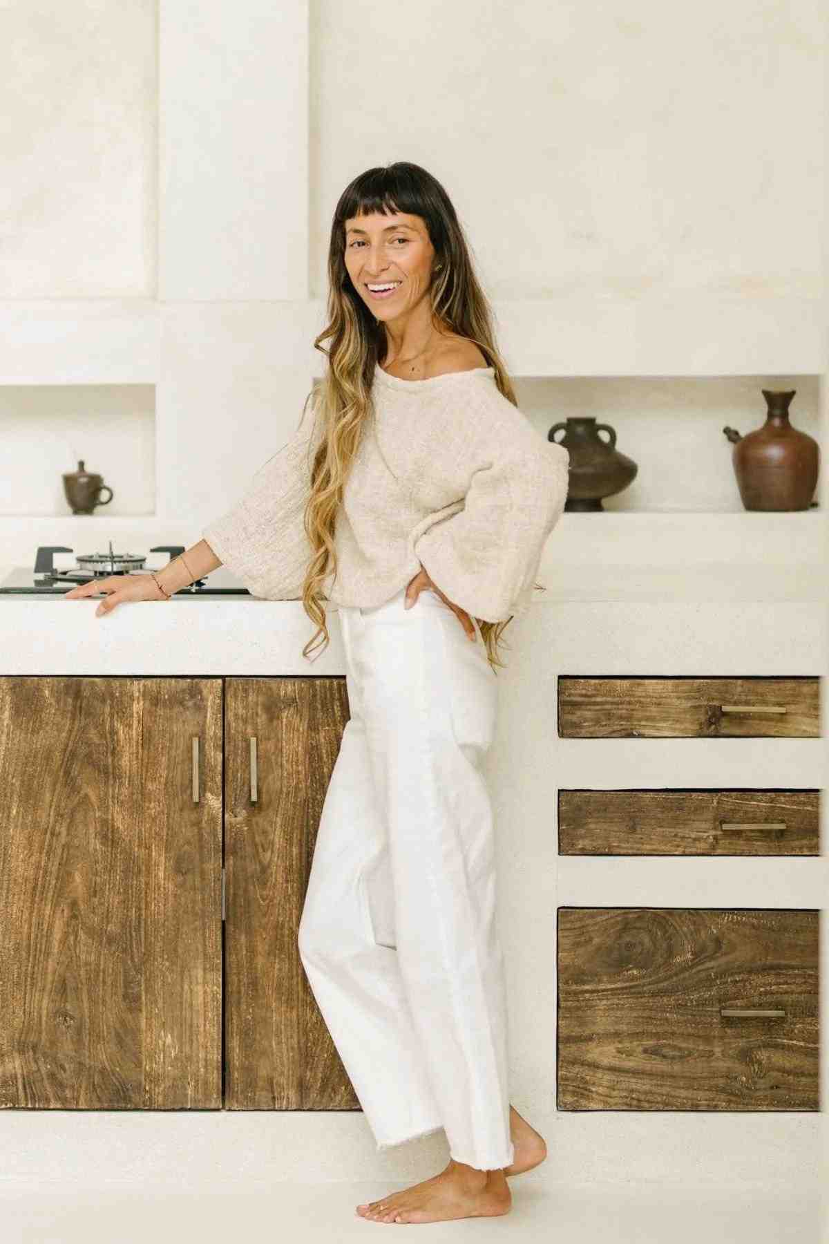 A relaxed woman with long hair stands barefoot in a minimalist kitchen, wearing an oversized beige Tumanggal Cotton Spirit Top by Myrah Penaloza and white pants. The kitchen features white countertops and wooden cabinets as she smiles slightly.