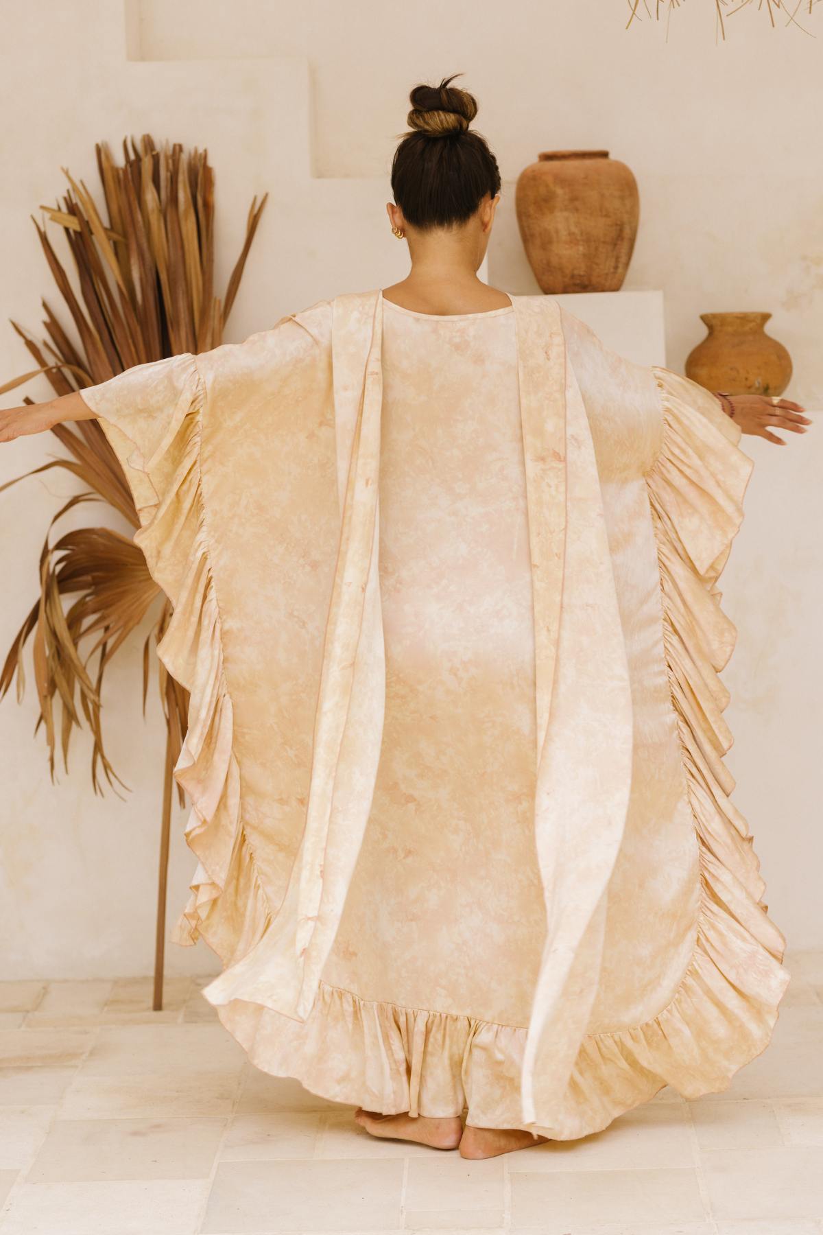 A person stands facing away from the camera with arms outstretched, showcasing the Myrah Penaloza Perla Silk Gown Made To Order, a flowing, ruffled beige dress that evokes Florence and The Machine's ethereal style. The background highlights dried plants and rustic clay pots arranged on shelves. The person's hair is elegantly tied up in a bun.