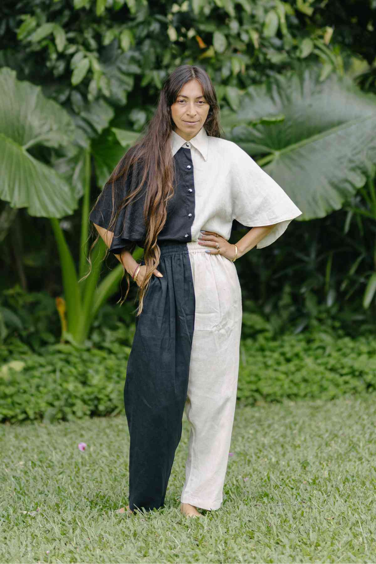 A woman with long hair stands on grass, dressed in a distinctive Linen Kiki Yin Yang Set (Made To Order) from Myrah Penaloza. The ensemble, crafted from 100% stone-washed linen, features a two-toned, short-sleeved shirt and coordinating pants. Behind her, lush greenery with large leaves creates a vibrant, natural backdrop.