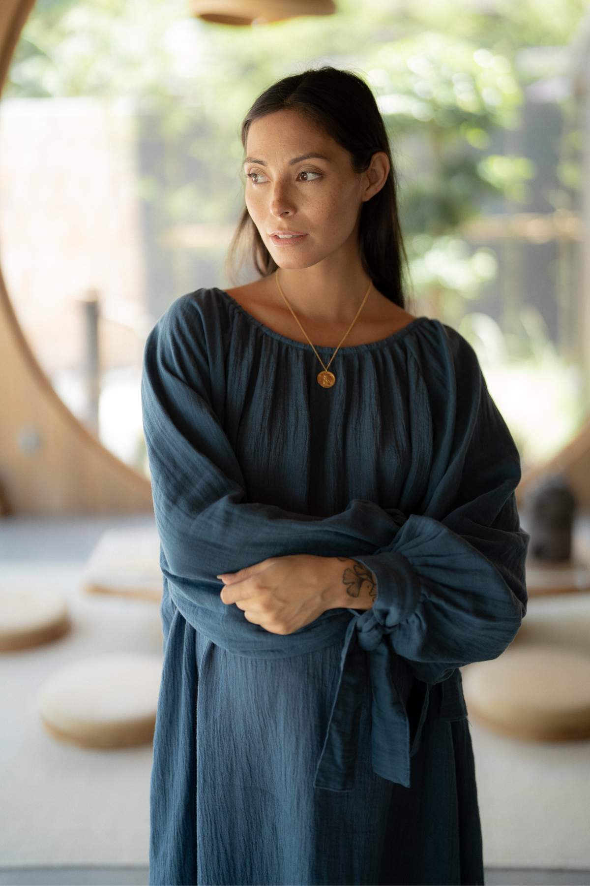 A woman in a Myrah Penaloza Longing Gown (reversible, BI) stands indoors under soft lighting. Her long dark hair cascades over a pendant necklace, while the circular wooden architecture in the background adds a rustic touch, complemented by blurred greenery that enhances the Virgo details of her look.