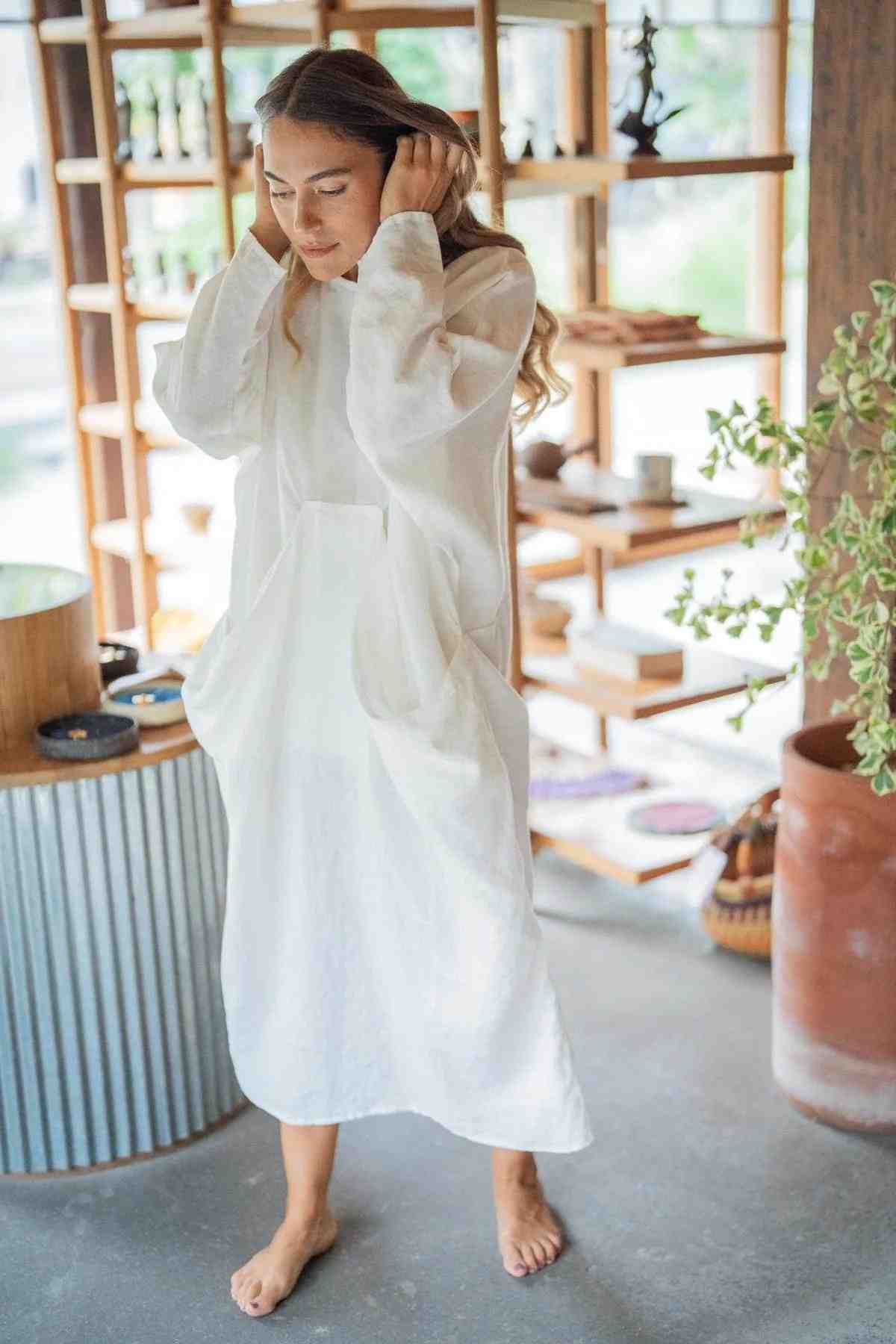 A woman with long hair stands barefoot in a bright room, adorned in a Mountain Magic Linen Gown by Myrah Penaloza. She has her hands raised to her head, the loose, flowing dress adding an air of elegance. Behind her is a wooden shelf with various objects and a large potted plant to her right.