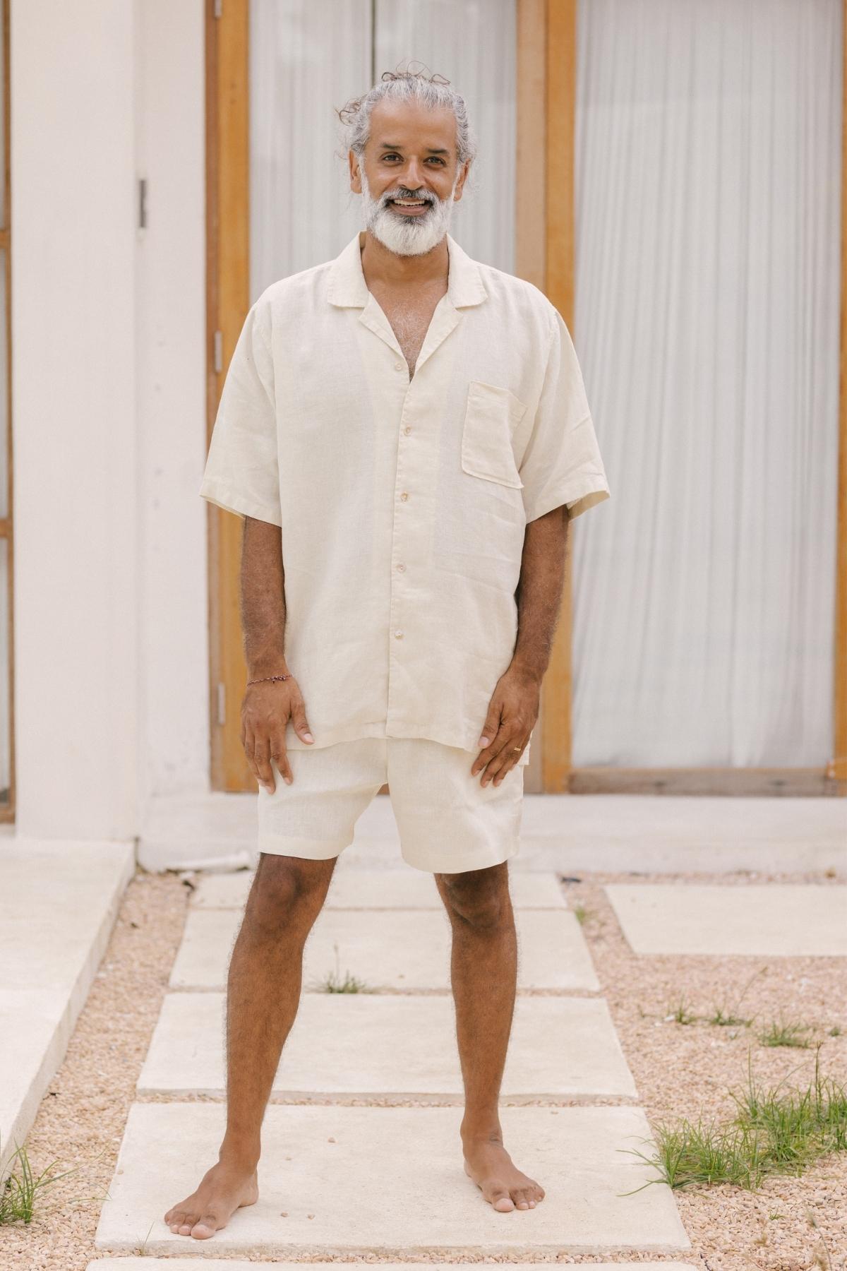 A barefoot man with gray hair and a beard, embodying the charm of a nomadic traveler, smiles while standing on a stone path. He is dressed in Myrah Penaloza's Caravana Men's Linen Top & Short. Two wooden doors with white curtains frame an inviting backdrop.