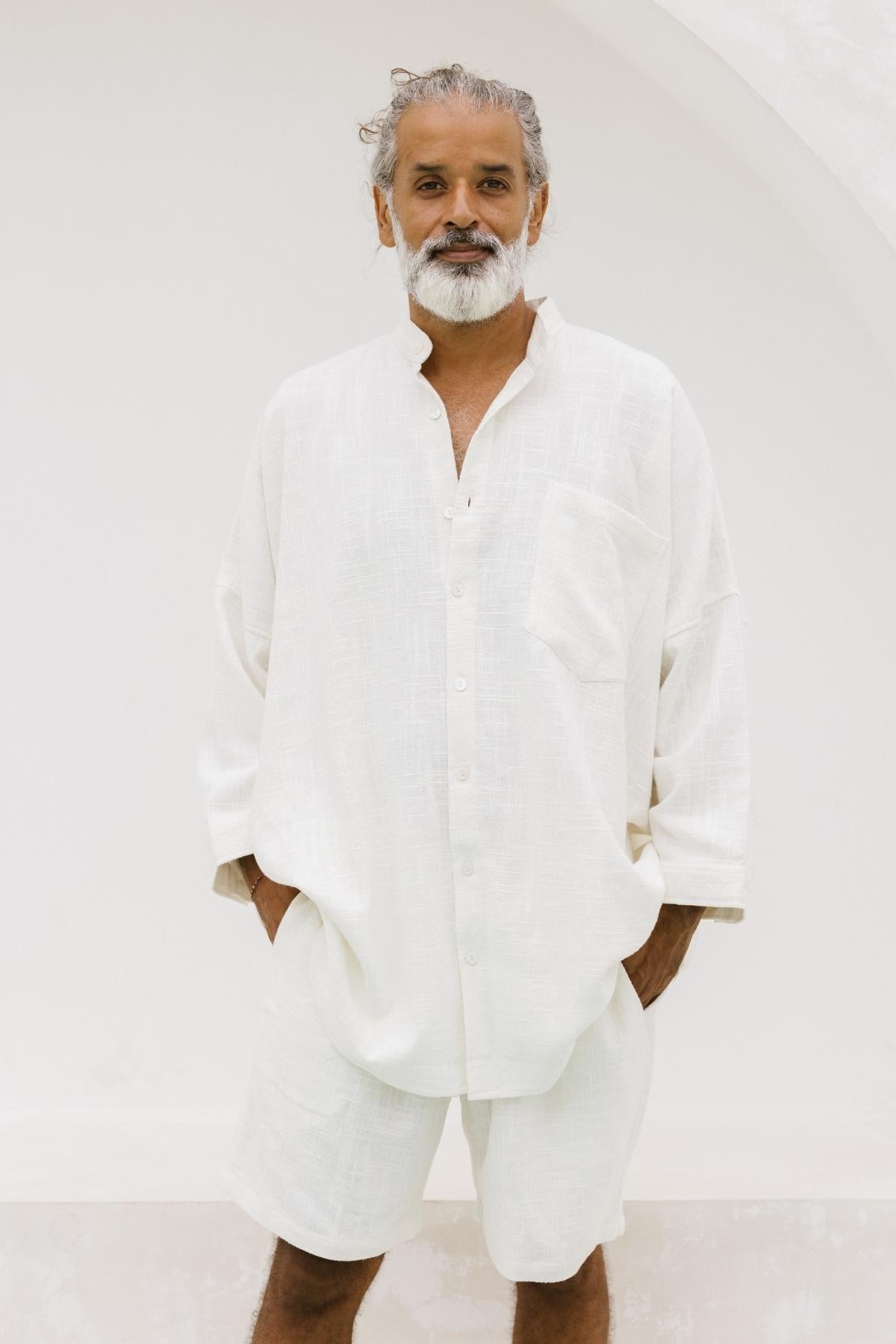 A man with gray hair and a beard stands against a white backdrop, wearing the Camisa Del Novio Mens by Myrah Penaloza. This off-white ensemble is crafted from linen and cotton, showcasing an oversized pocket while he casually rests his hands in his pockets.