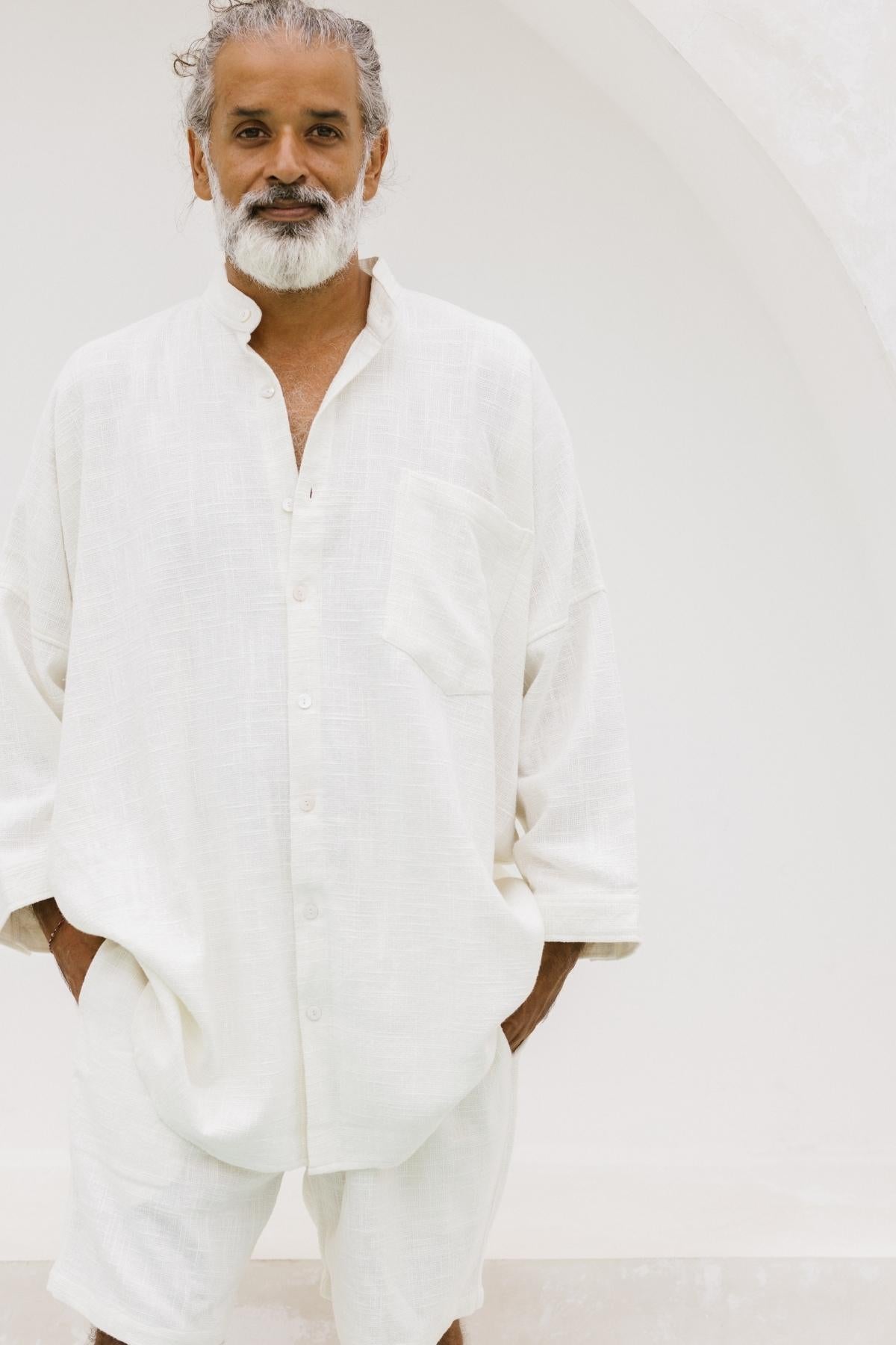 An individual with gray hair and a beard stands against a white background, exuding a calm and relaxed demeanor. They are dressed in the loose-fitting Camisa Del Novio Mens by Myrah Penaloza, an off-white linen/cotton shirt and shorts ensemble made to order, featuring oversized pockets.