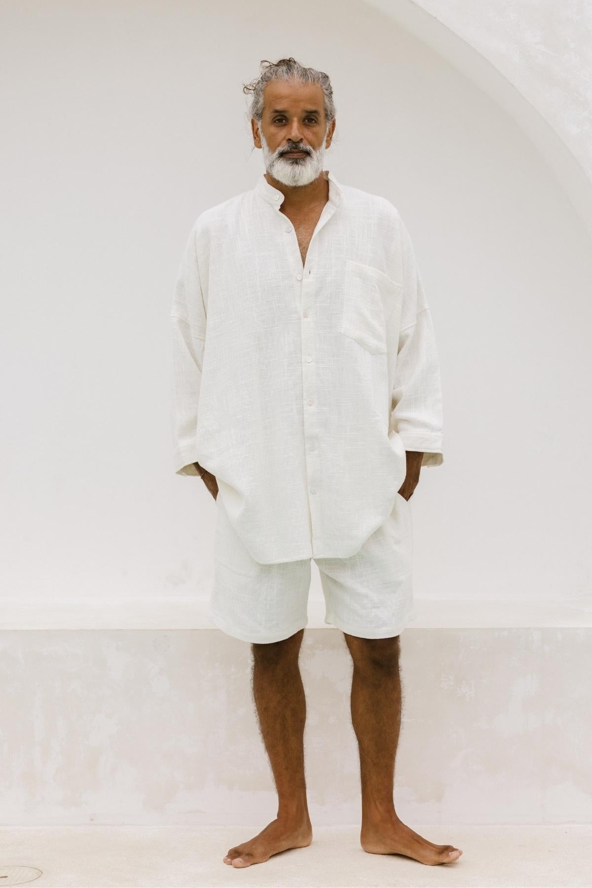 A man with a beard stands barefoot against a light-colored archway, wearing Myrah Penaloza's Camisa Del Novio Mens ensemble in off white. Crafted from linen and cotton and made to order, his loose-fitting shirt with an oversized pocket and matching shorts beautifully complement his curly, graying hair.