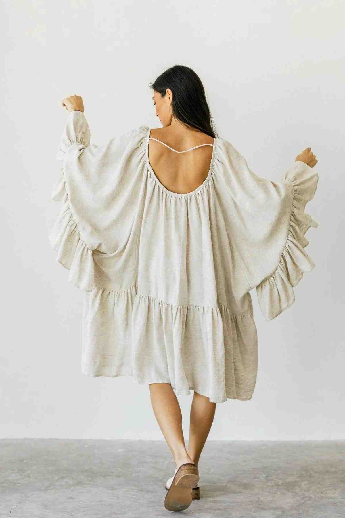 A person with long dark hair stands facing away, wearing a loose, flowy cream Linen Brida Gown Short by Myrah Penaloza. The romantic dress, ideal for spring or summer, showcases ruffled tiers and is beautifully made from linen fabric. They stand on a plain gray floor.