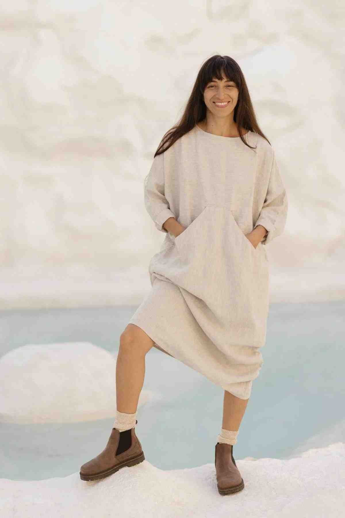 A woman with long dark hair, wearing a loose beige Mountain Magic Linen Gown by Myrah Penaloza and brown ankle boots, is standing outdoors with one leg slightly raised on a rock. She is smiling and has her hands in her dress pockets. The background is a serene, light-colored natural setting.