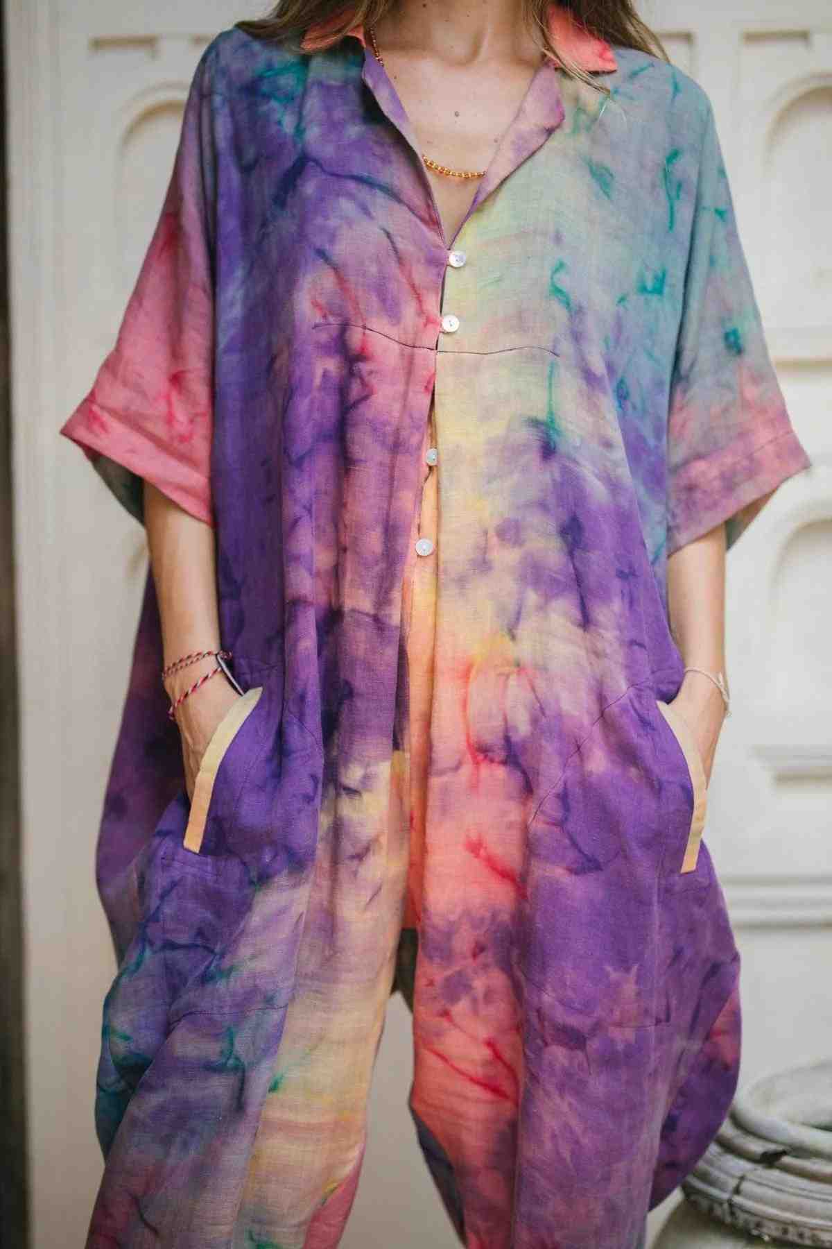 A person wearing the vibrant Sat Torri Rainbeau Playsuit by Myrah Penaloza, showcasing a retro 1980's tie-dye design in shades of purple, pink, green, and yellow, stands with hands in their pockets. The playsuit features buttons down the front and short sleeves. The background is a light-colored textured wall.