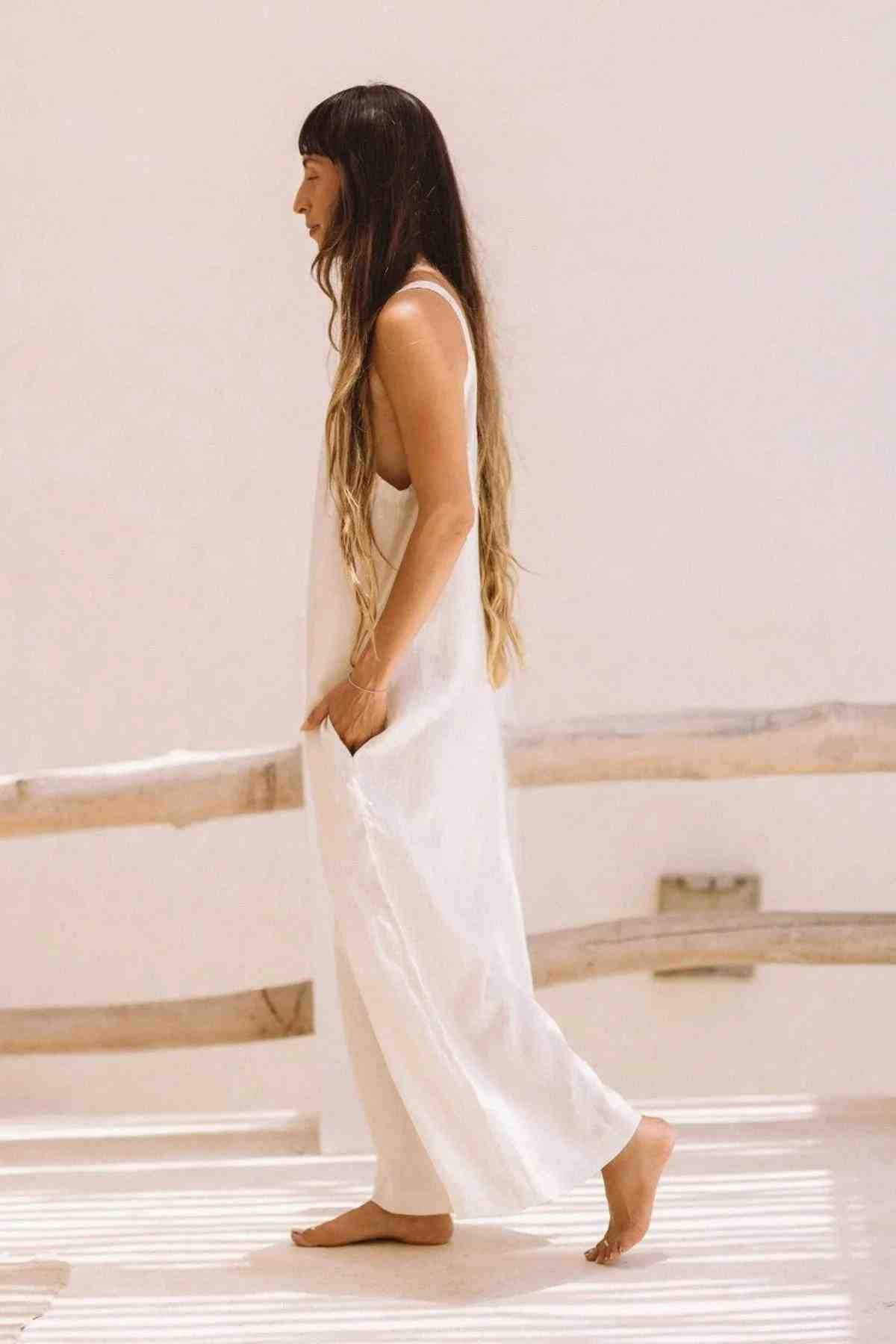 A person with long hair, wearing the Kundalini Linen Playsuit by Myrah Penaloza, crafted from luxuriously soft stonewashed linen, walks barefoot indoors. The background is bright and minimal, featuring light-colored wooden elements.