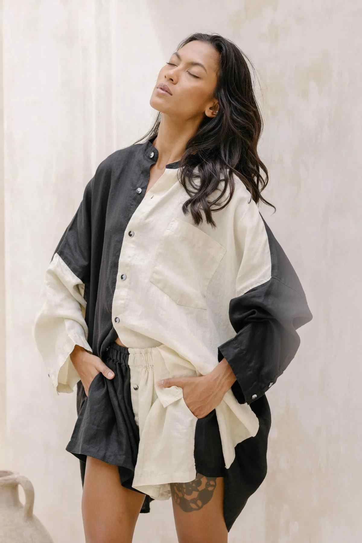 A person stands against a plain light background, wearing the Unisex Yin Yang Linen Camisa Del Novio Set by Myrah Penaloza. The set features a loose-fitting, long-sleeved, color-blocked shirt in black and cream adorned with a Yin Yang linen patch, paired with matching shorts. They have long, wavy hair and their eyes are closed, with hands placed in their pockets.