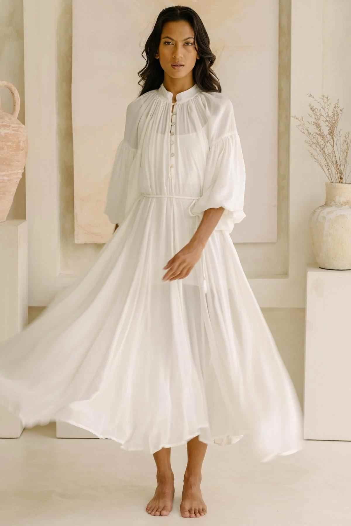 A woman stands barefoot in Myrah Penaloza's bridal-inspired Paloma Silk Gown, a luxurious white dress with long, puffed sleeves and button details on the bodice. She stands against a minimalistic beige background adorned with two large pottery vases and abstract wall art.
