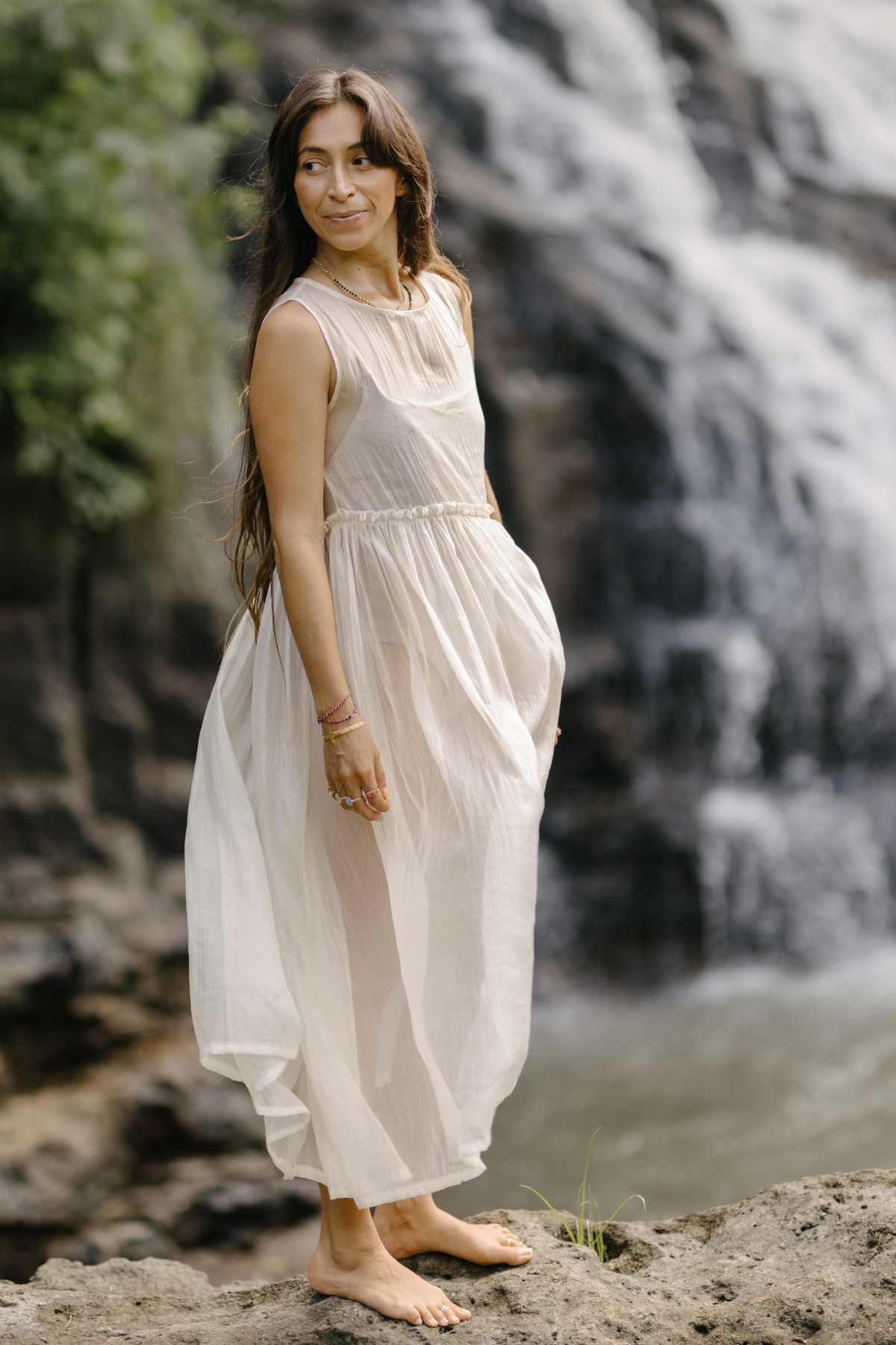A woman stands barefoot on a rocky surface near a waterfall, wearing the Dolly Night Gown by Myrah Penaloza — a sleeveless, sheer design made from 100% chiffon silk. With her long brown hair cascading down her back, she gazes to the side with a serene expression amidst lush greenery and the backdrop of cascading water.
