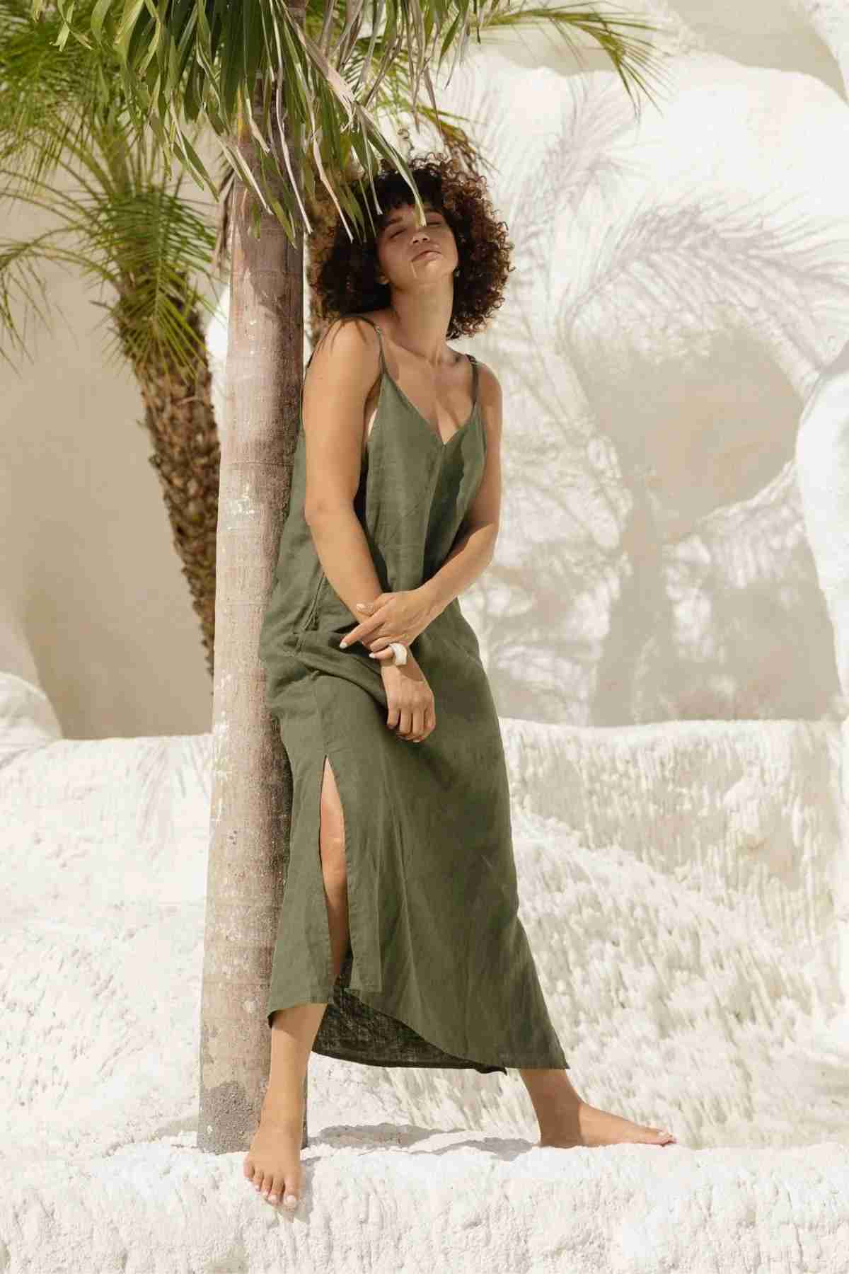 A barefoot woman in a green dress, reminiscent of Myrah Penaloza's Everyday Linen Slip, stands leaning against a palm tree, surrounded by white sand. She is outdoors in a sunny environment, with her eyes closed and head tilted back, exuding luxurious comfort and a serene vibe.