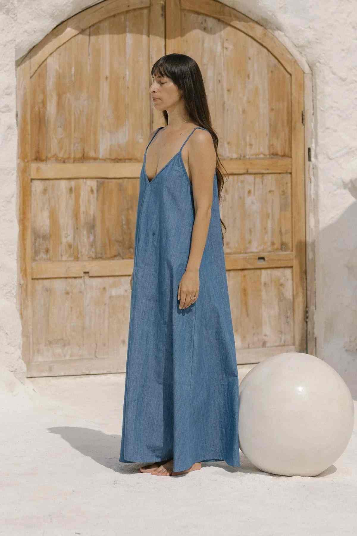 A woman stands barefoot on a sandy surface, wearing the loose-fitting Kundalini Denim Dress by Myrah Penaloza. With thin straps and stonewashed linen, the maxi dress complements her long dark hair as she poses in front of a large wooden door set in a white wall. To her right, there is a large white spherical object.