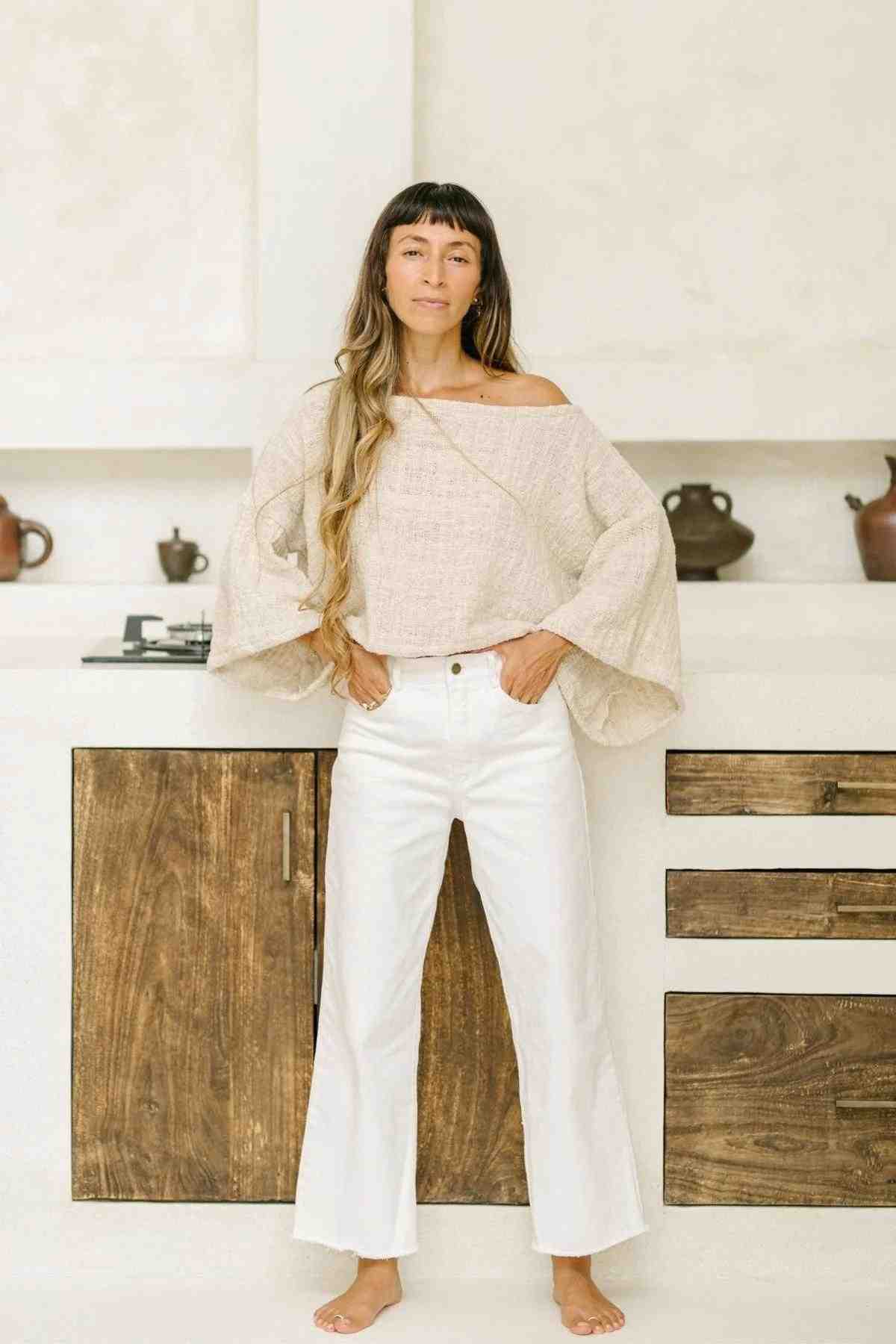 A woman with long hair stands barefoot in a kitchen, wearing the Tumanggal Cotton Spirit Top from Myrah Penaloza—a loose, off-the-shoulder beige top made of hand-woven cotton—and white pants. The kitchen features wooden cabinets and a countertop adorned with a few pots and decorations. She has a calm expression and stands with her hands in her pockets.
