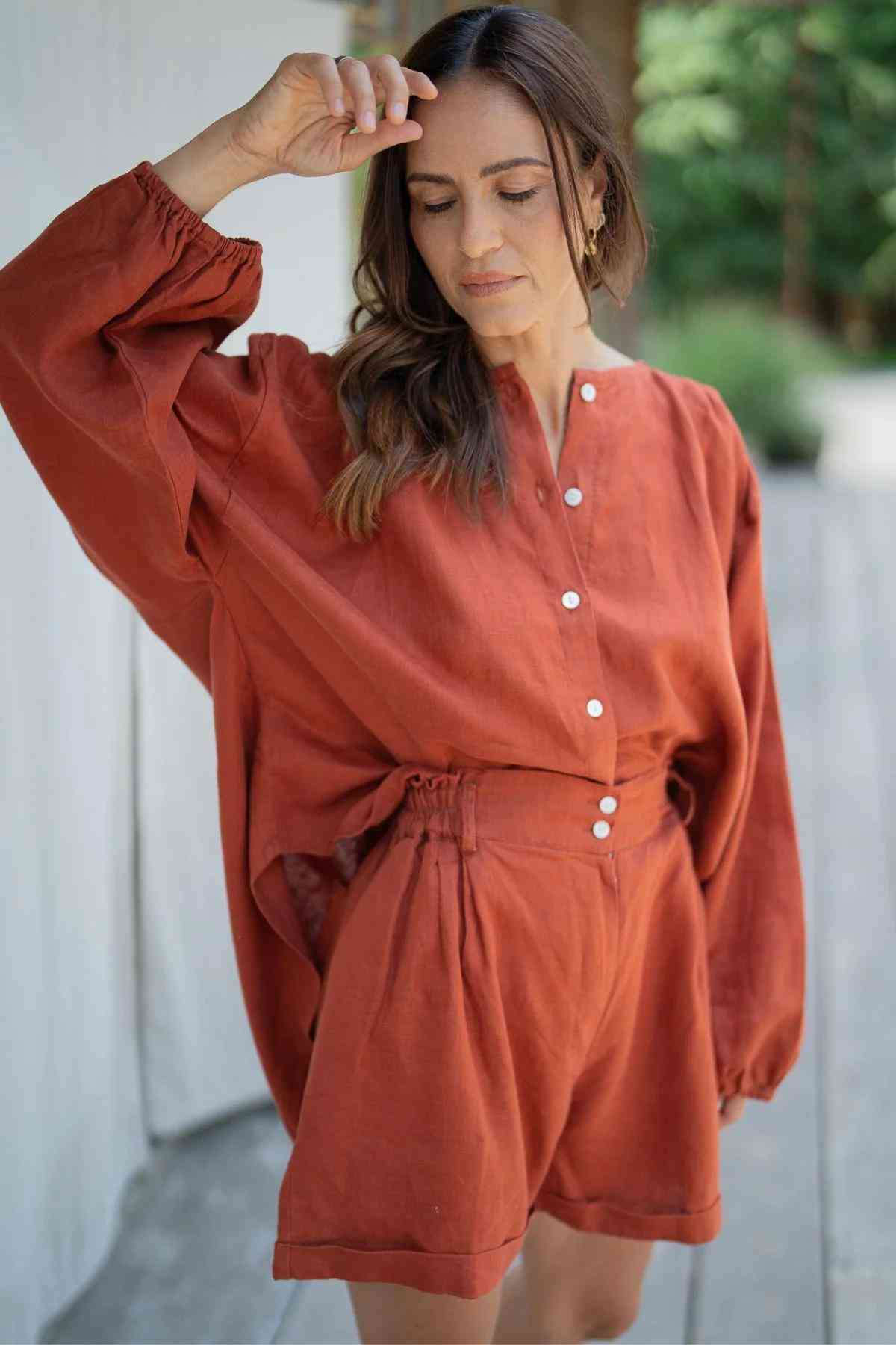 A woman stands outdoors in an elegant stone-washed linen ensemble, featuring the Rust Rice Linen Set from Myrah Penaloza. She raises her left arm, touching her forehead with her hand. The blouse showcases billowy sleeves and buttons down the front. The background is blurred with greenery and a wooden surface.