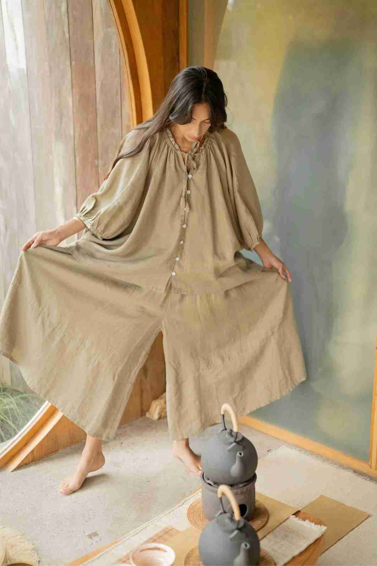 A person wearing the wide-legged Pantalon Linen Pants by Myrah Penaloza poses indoors near a large round window with a wooden frame. Decorative pots are placed on the floor in the foreground, and soft natural light fills the space.