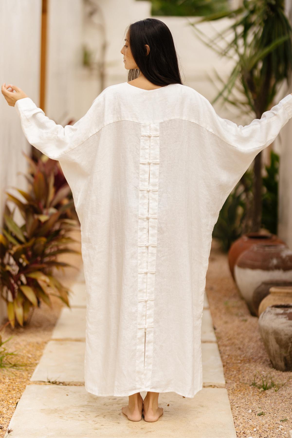 A person with long dark hair is standing outside with their back to the camera, arms slightly raised, wearing a loose-fitting, long white garment adorned with Japanese-inspired buttons - the Ancient Chajin Jacket (100% Linen) by Myrah Penaloza. They are in a garden area, surrounded by plants and large clay pots next to a stone path.