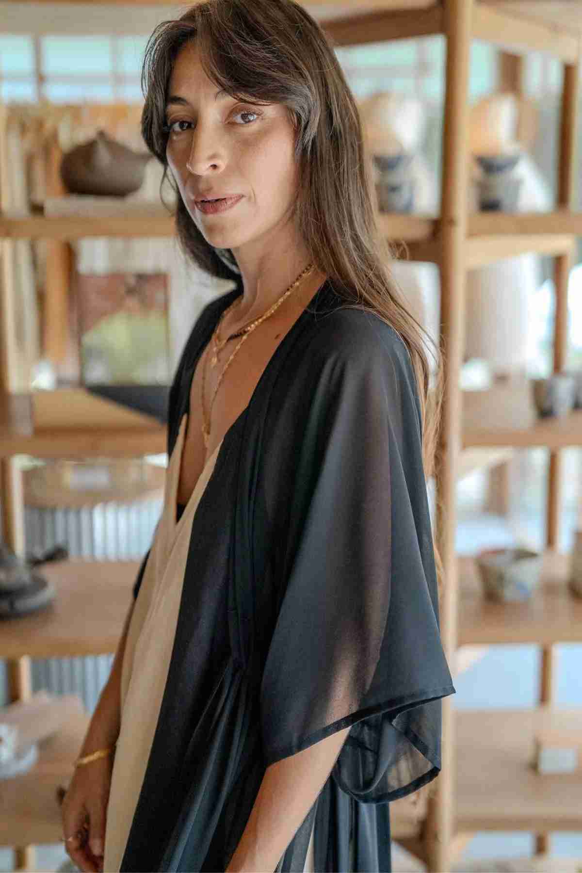 A woman with long brown hair and a gentle smile stands in front of wooden shelves displaying various items. She is wearing the Madelin Sheer Chiffon Long Cover by Myrah Penaloza over a white dress, along with several necklaces. The setting appears to be a cozy, well-lit shop or studio.