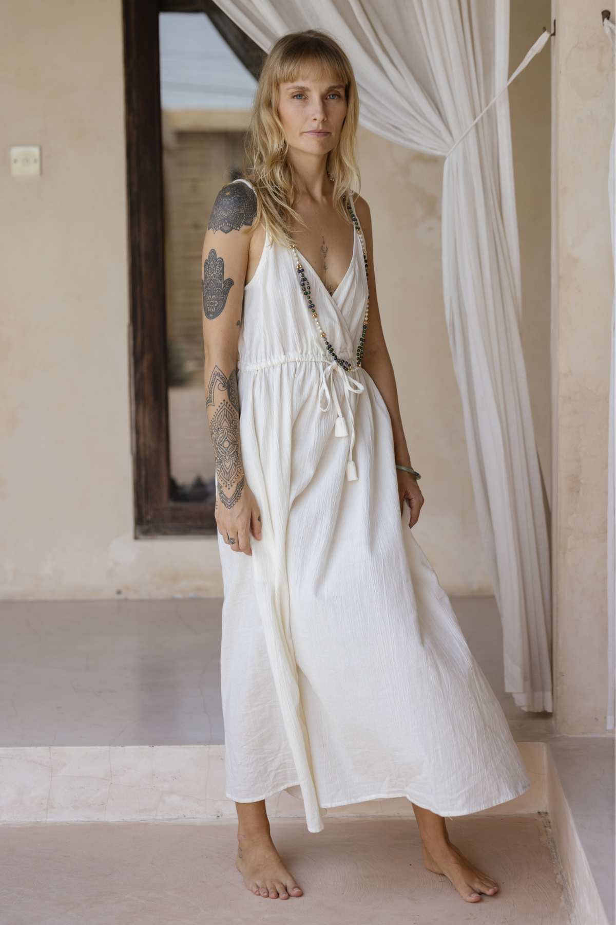 A person with long hair stands barefoot on a tile floor, exuding effortless elegance in a sleeveless white Dora Slip Dress by Myrah Penaloza, complete with a tie at the waist. Their tattooed arms add character, while a sheer curtain and wooden-framed door provide the perfect backdrop for this timeless piece.