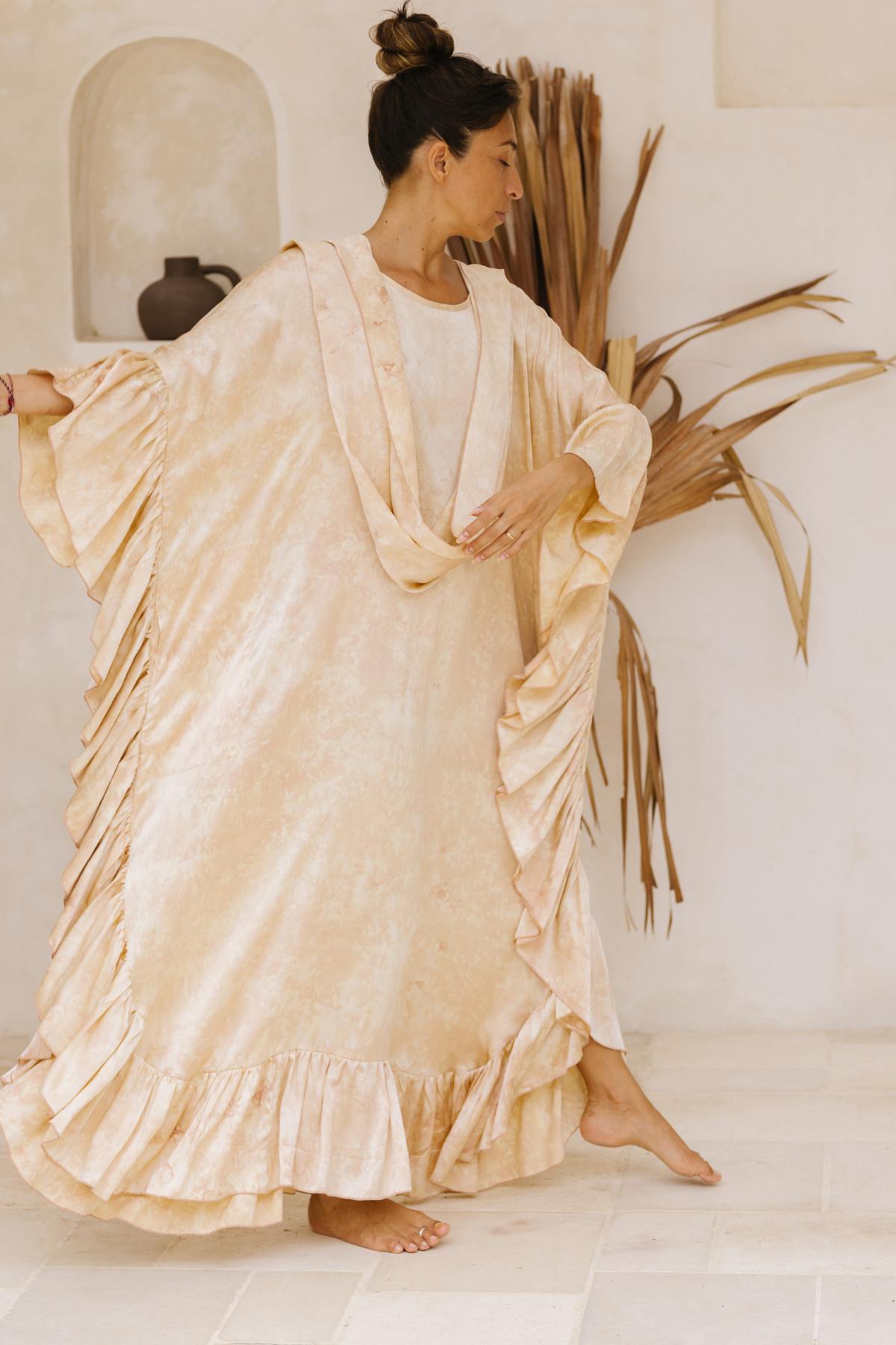 A woman stands barefoot indoors, striking a graceful pose while wearing the Perla Silk Gown Made To Order by Myrah Penaloza. The flowing, beige dress with ruffled edges resembles a vintage wedding gown. Her hair is tied up in a bun, and the background features minimalistic decor, including a vase and dried palm leaves against a light-colored wall.