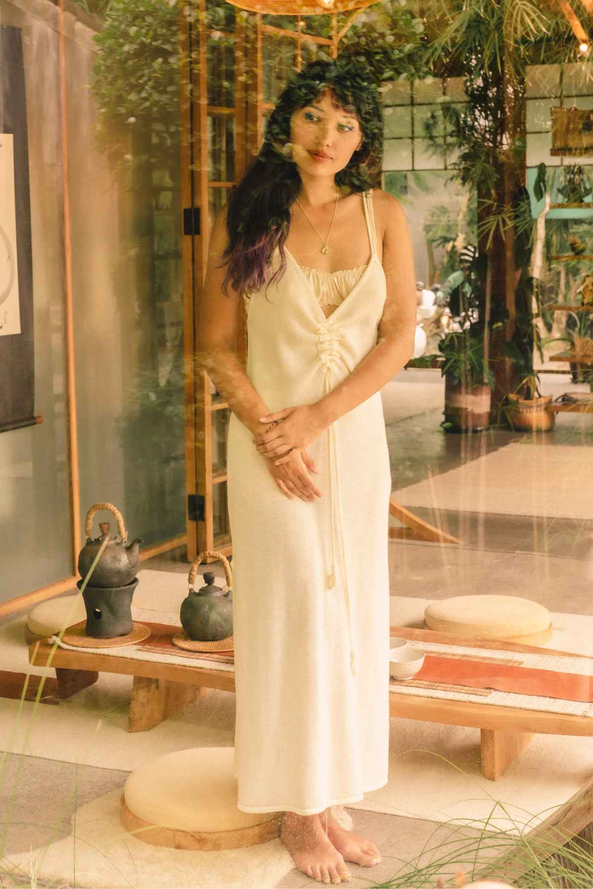 A woman stands barefoot in front of a glass door, wearing the Virgo Slip Hand Knit Cotton by Myrah Penaloza. The setting includes wooden furniture and greenery, creating a warm, serene atmosphere.