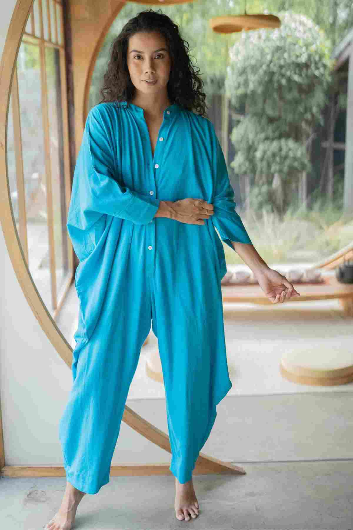 A woman with long, curly hair stands barefoot indoors near a circular wooden doorway. She is wearing a bright blue, loose-fitting Miracle Playsuit 2.0 from the Myrah Penaloza Karama Collection - PR. One hand is adjusting her sleeve, and the background reveals plants and a serene interior.