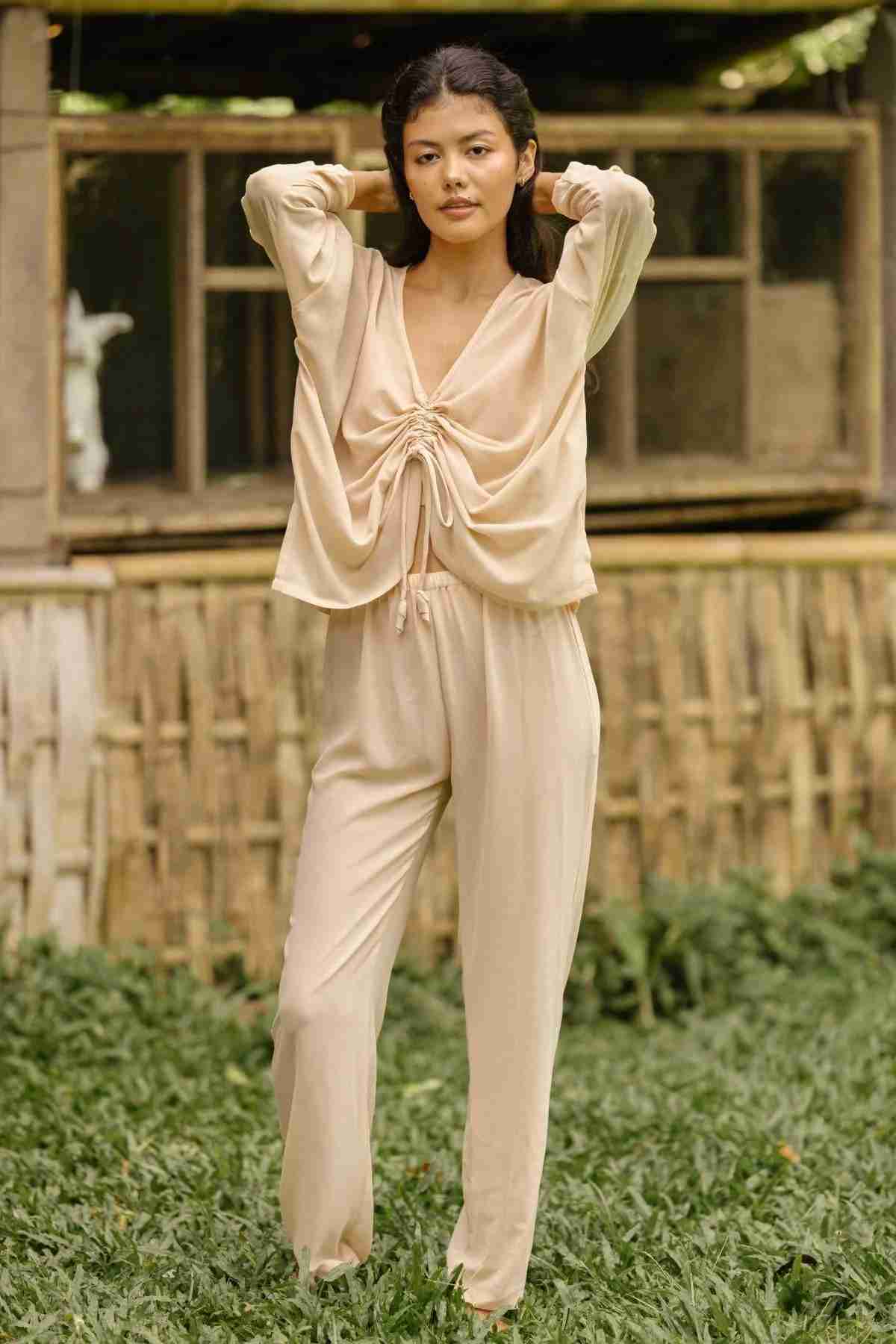 A person standing on grass in front of a rustic wooden structure is wearing the Chiffon Silk Sheer Virgo Rising Set (Made To Order) from Myrah Penaloza. Their hands are resting behind their head, and they have a calm expression.