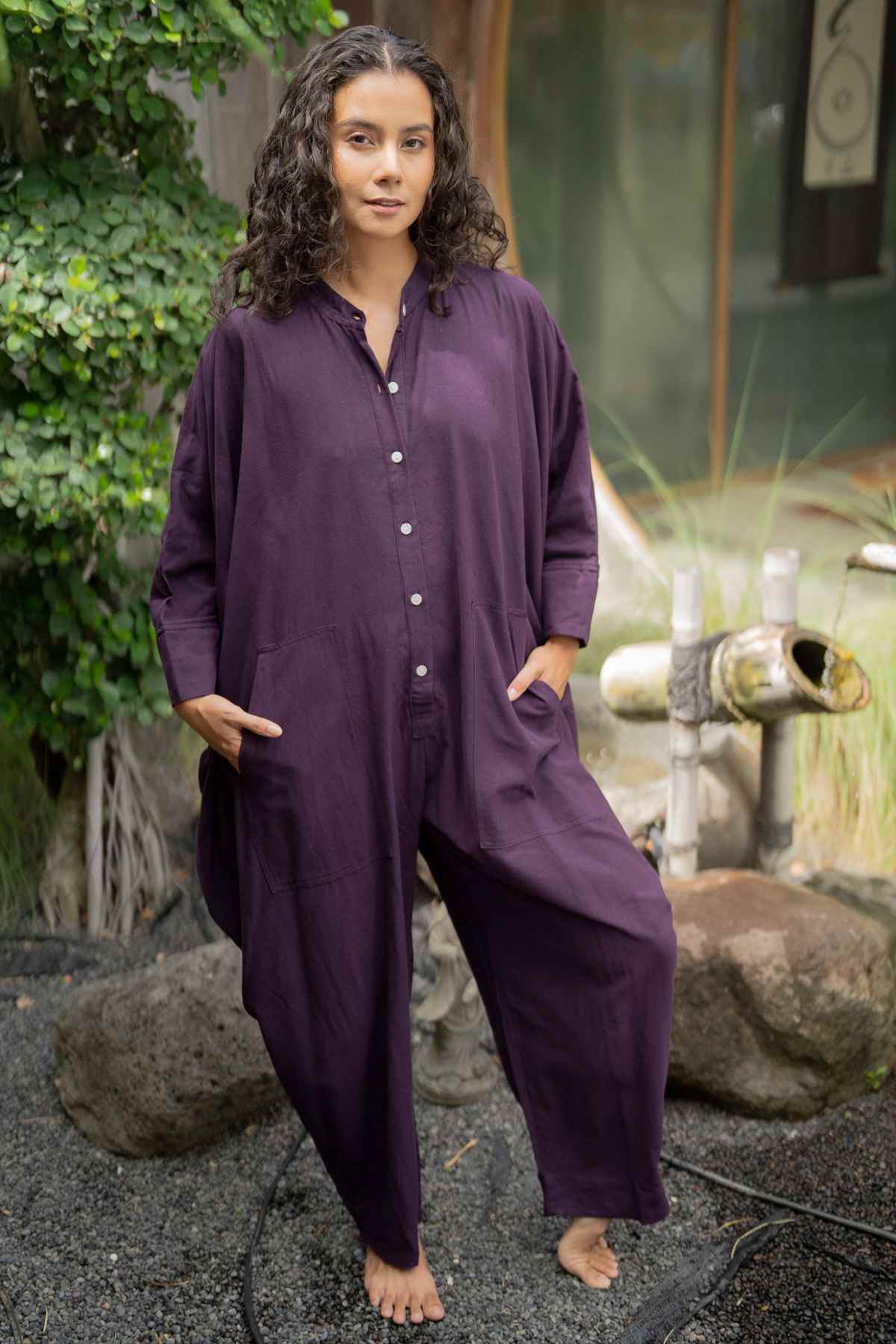 A woman with curly hair stands barefoot on a gravel surface outdoors, wearing a loose-fitting, dark purple Miracle Playsuit 2.0 from the Karama Collection by Myrah Penaloza, featuring buttons down the front and gazing at the camera. The backdrop showcases greenery, rocks, and a bamboo water feature, emphasizing this limited edition piece from our collaboration collection.